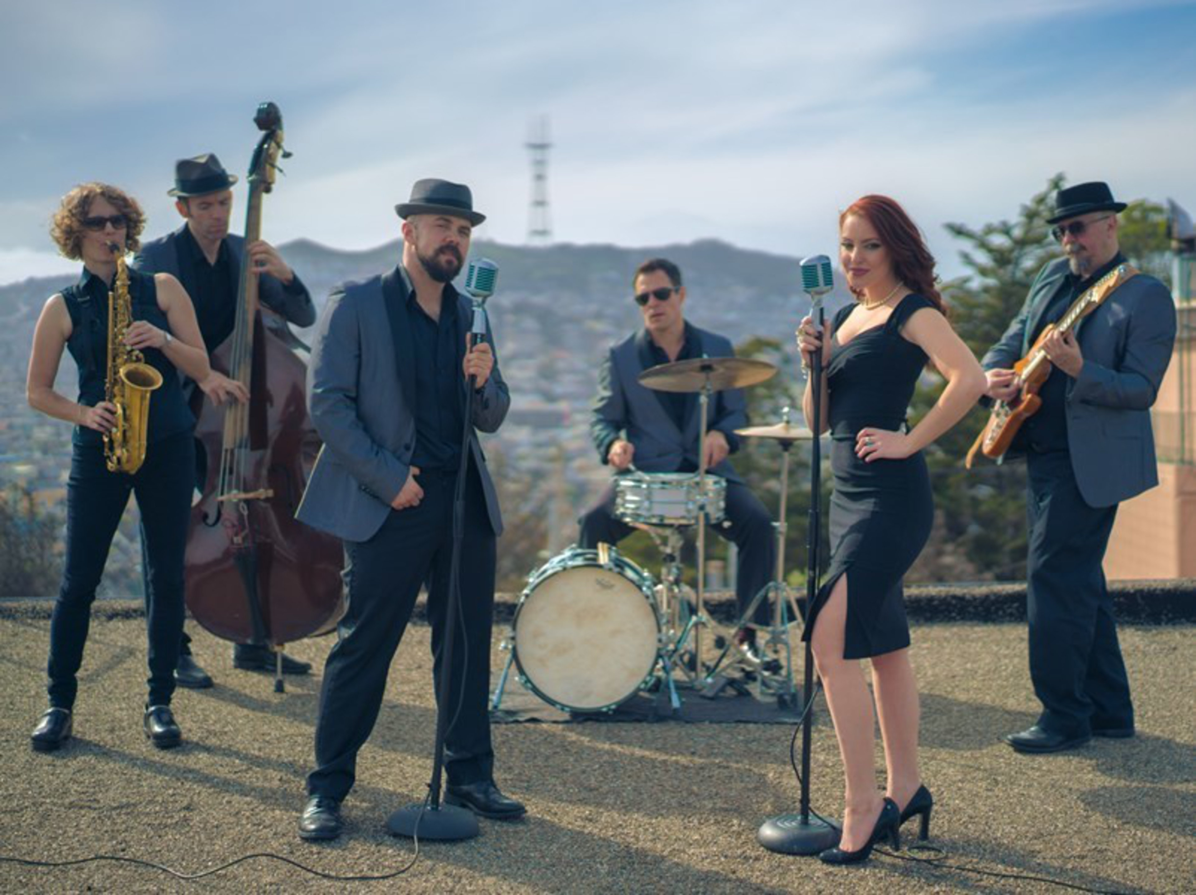 A six-member band performs outdoors with two singers at vintage microphones, accompanied by saxophone, upright bass, drums, and electric guitar.