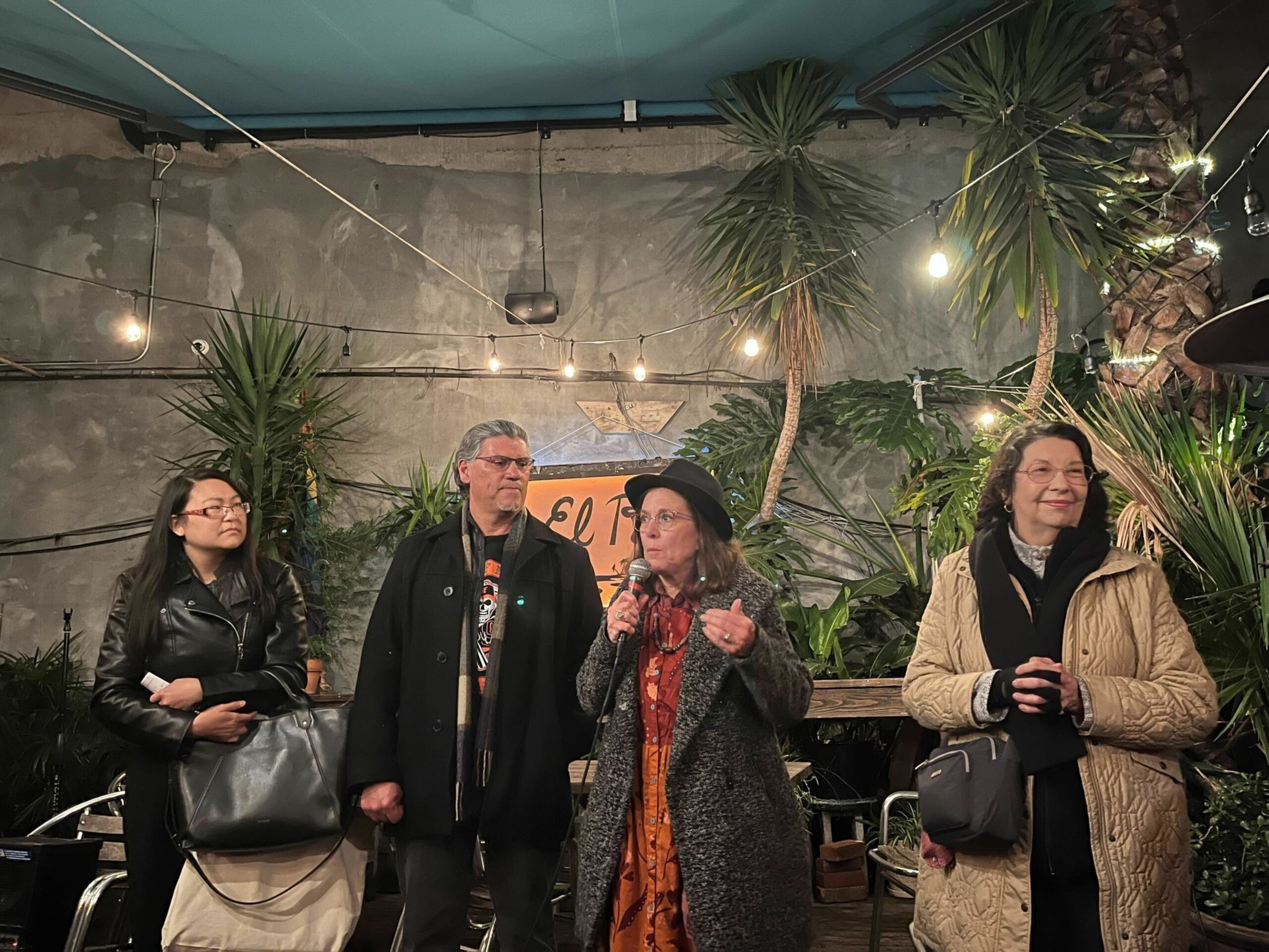 Four people stand in a warmly lit outdoor space with plants and string lights, one woman in a hat speaking into a microphone.