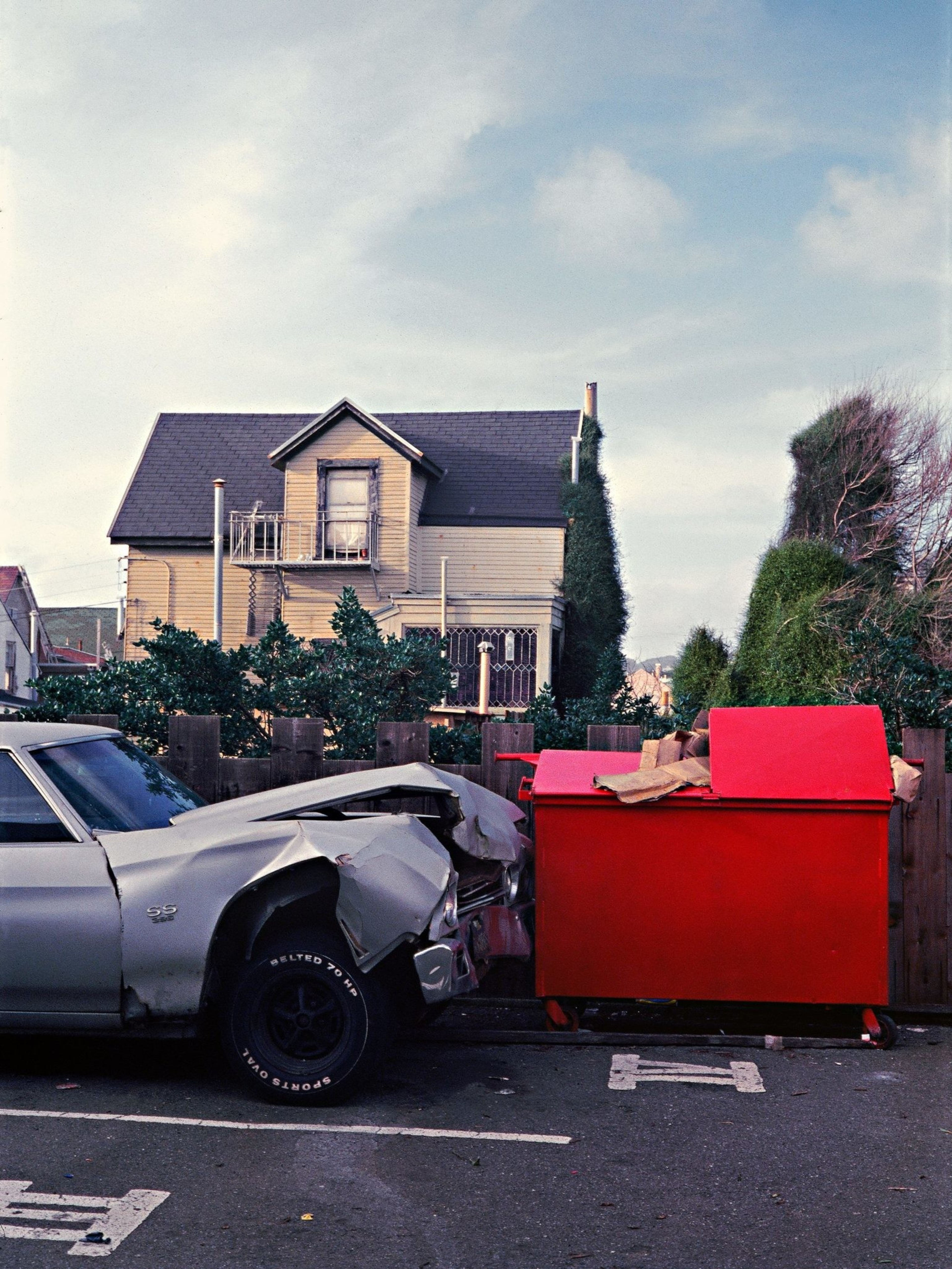 A damaged silver car is hitched to a bright red dumpster in a parking lot. Behind is a two-story house with a peaked roof, surrounded by trees and bushes.