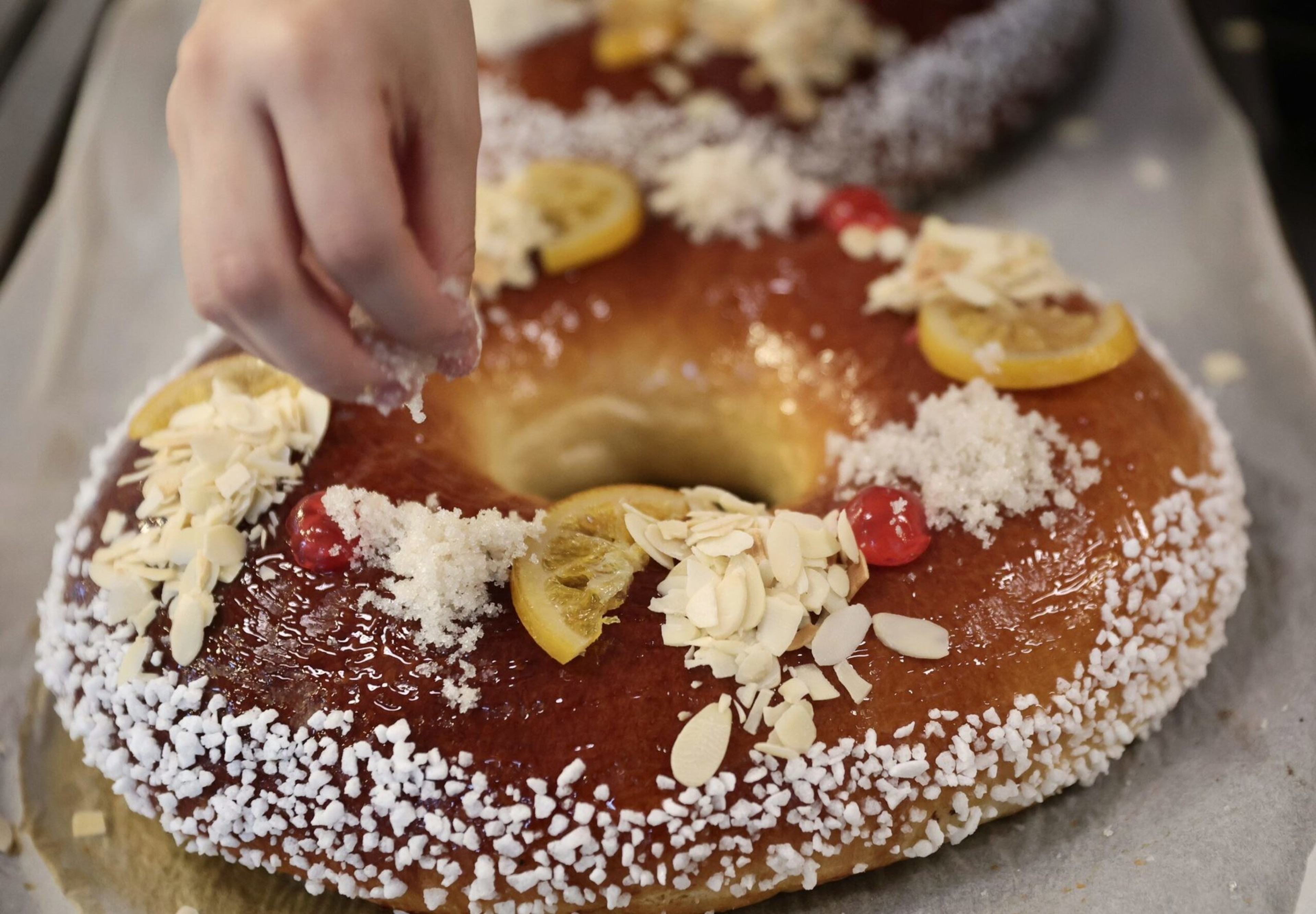 A hand is sprinkling sugar over a ring-shaped cake decorated with candied lemon slices, red cherries, almond flakes, and coarse sugar.