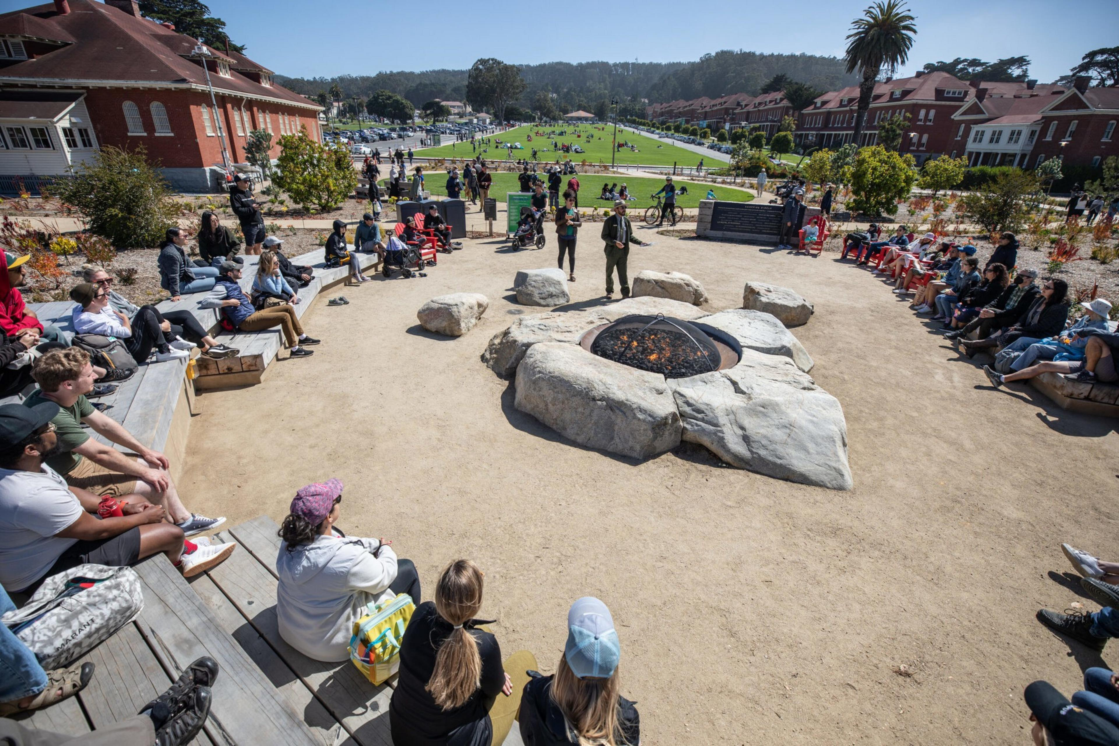 People sit around a large outdoor fire pit made of rocks, arranged in a circle, with buildings and green lawns in the background on a sunny day.