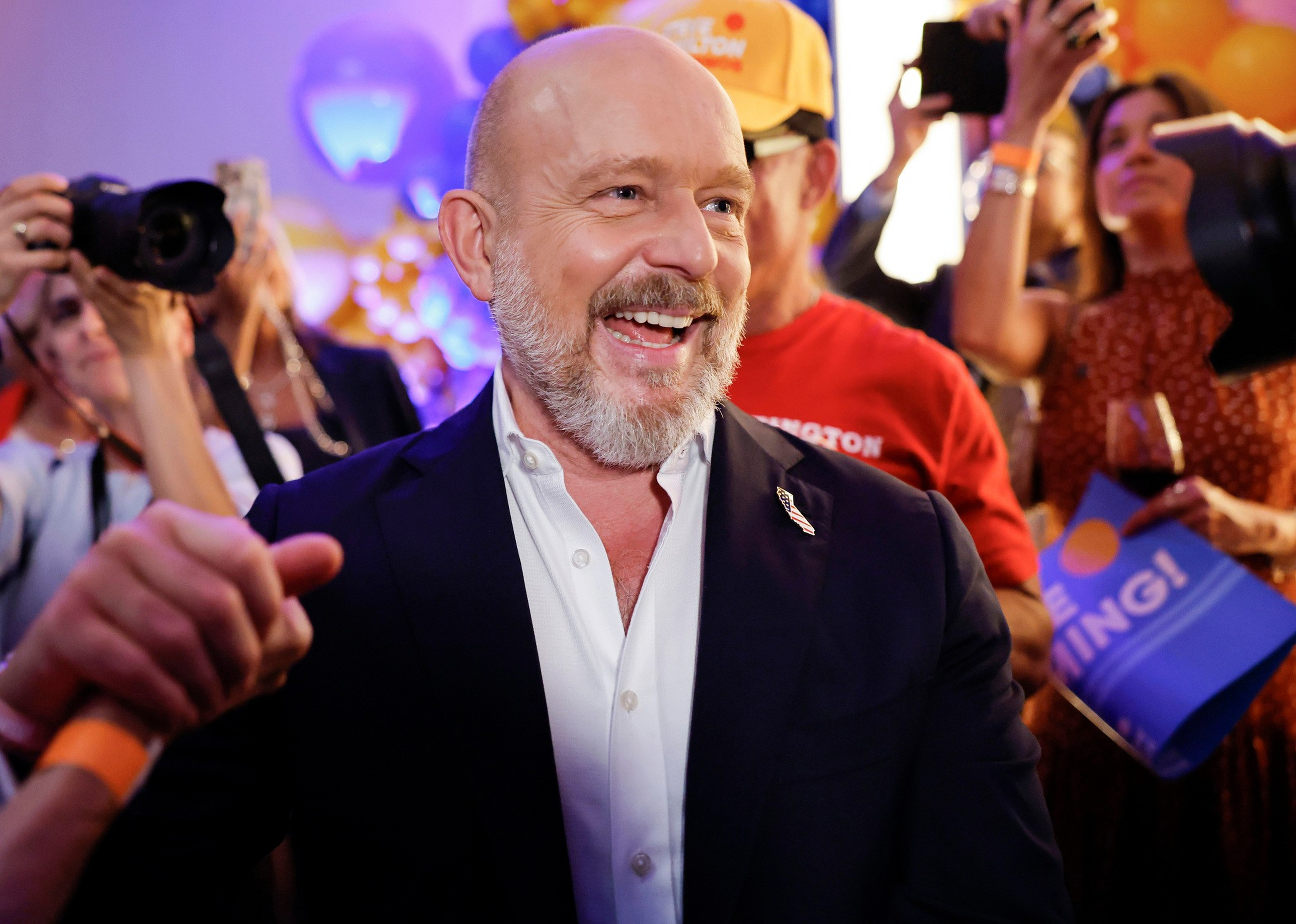 A smiling bald man with a beard wearing a dark blazer and white shirt shakes hands while people in the background take photos and hold signs.