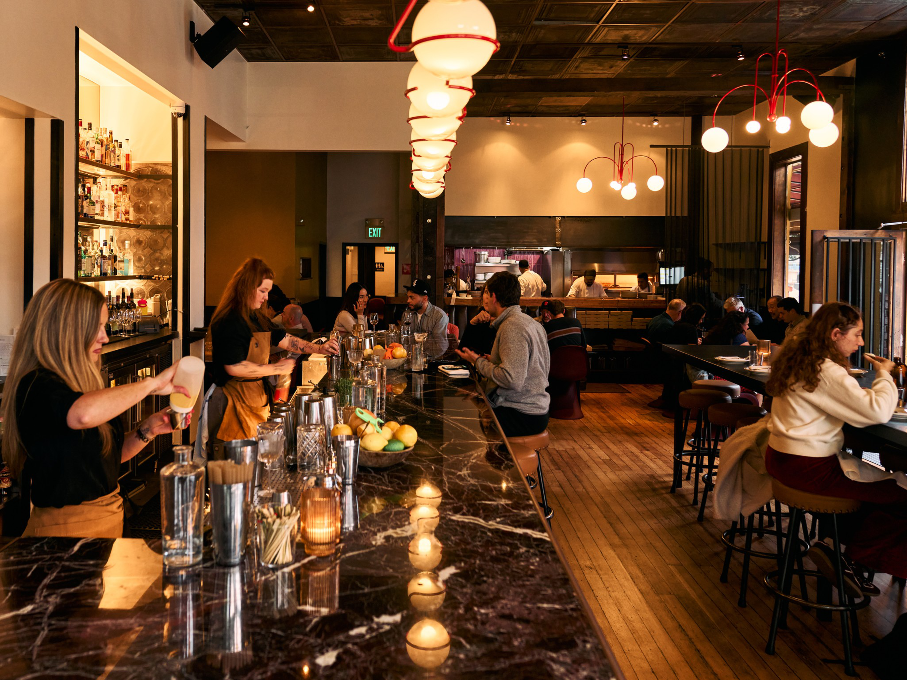 A warmly lit bar with patrons seated on stools, bartenders preparing drinks, and a cozy dining area in the background with wooden floors and modern lighting.