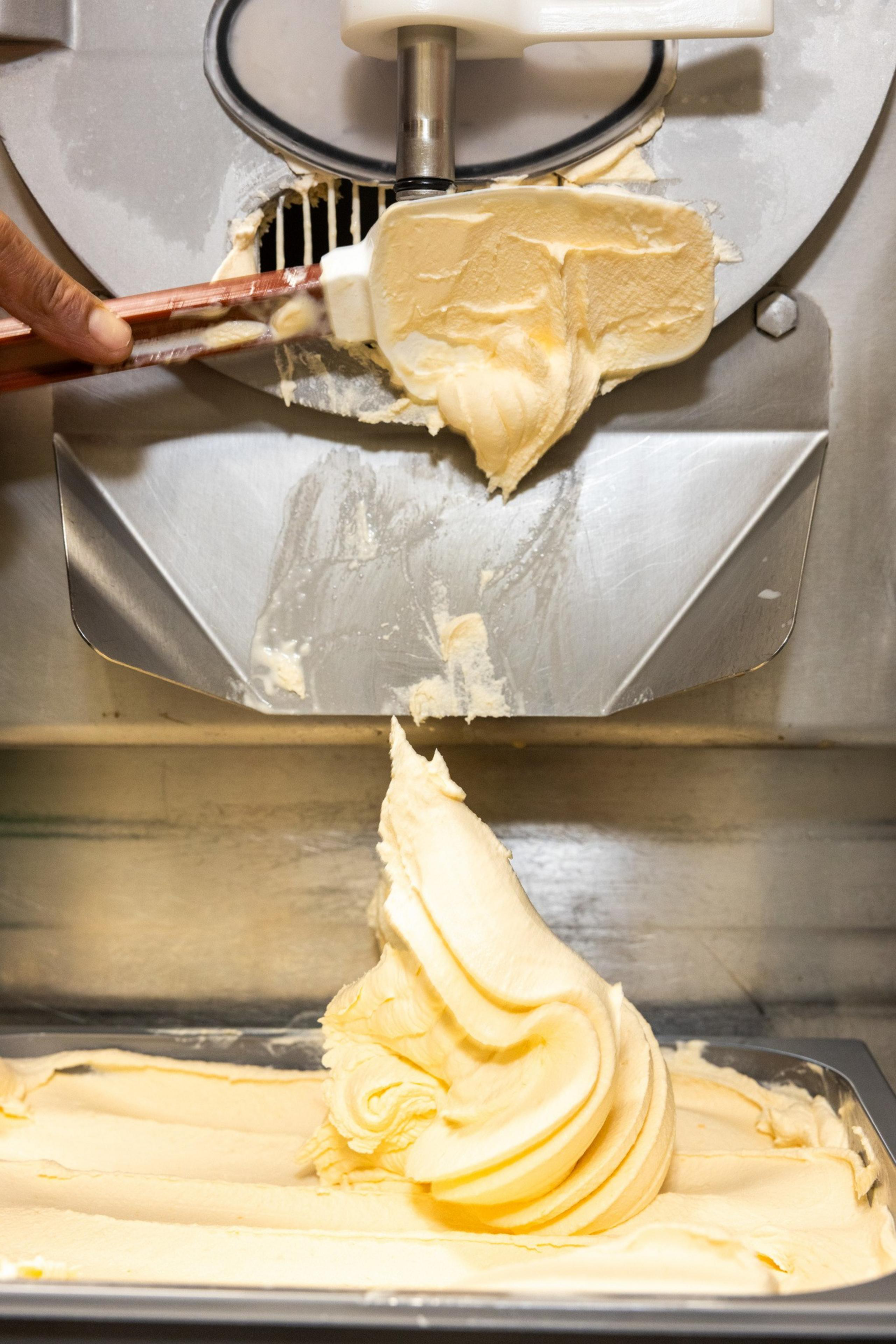 The image shows soft, yellow ice cream being dispensed from a metal machine into a container, with a hand using a spatula to guide the ice cream.
