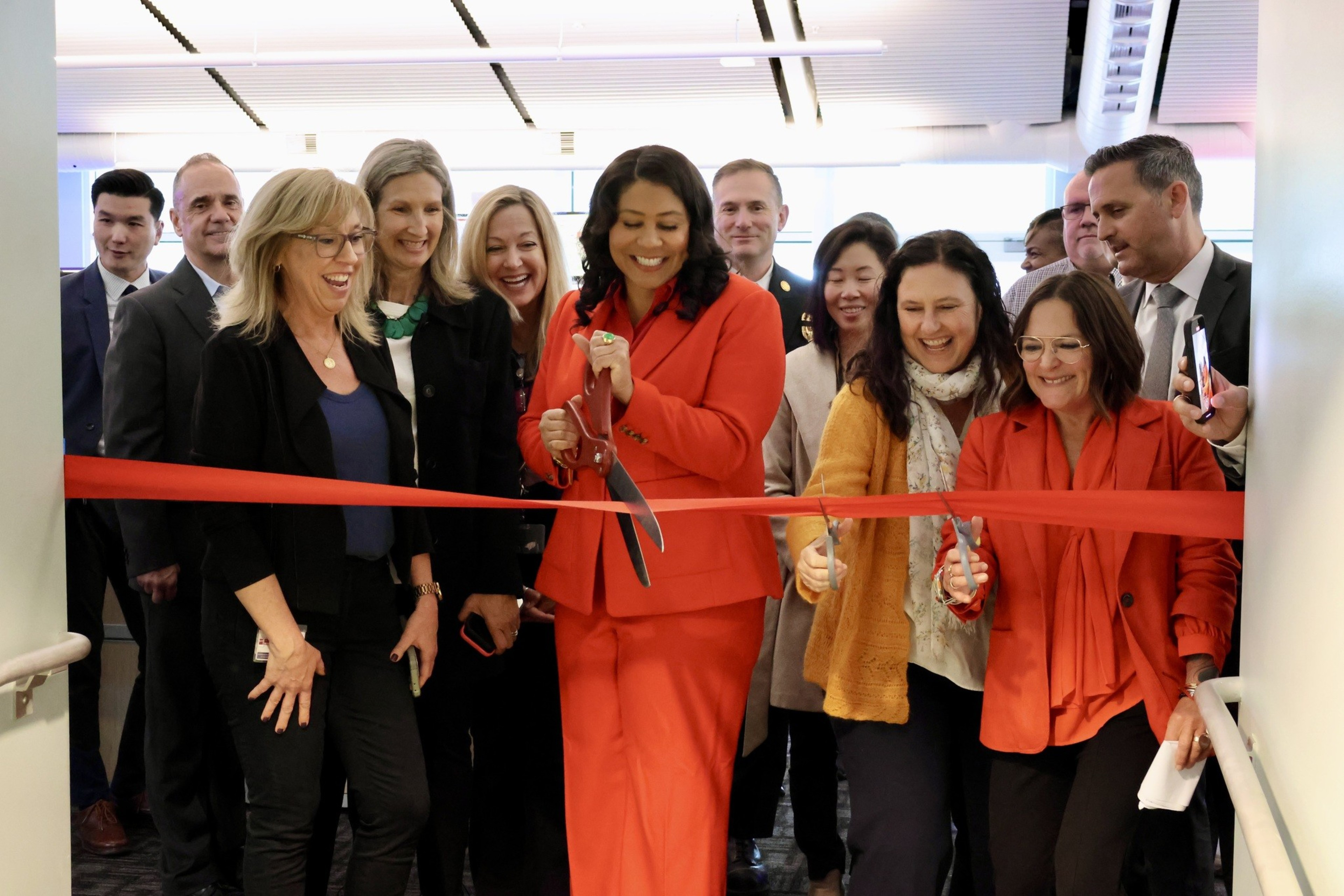 A group of smiling people watching a woman in a red suit cutting a ceremonial red ribbon.