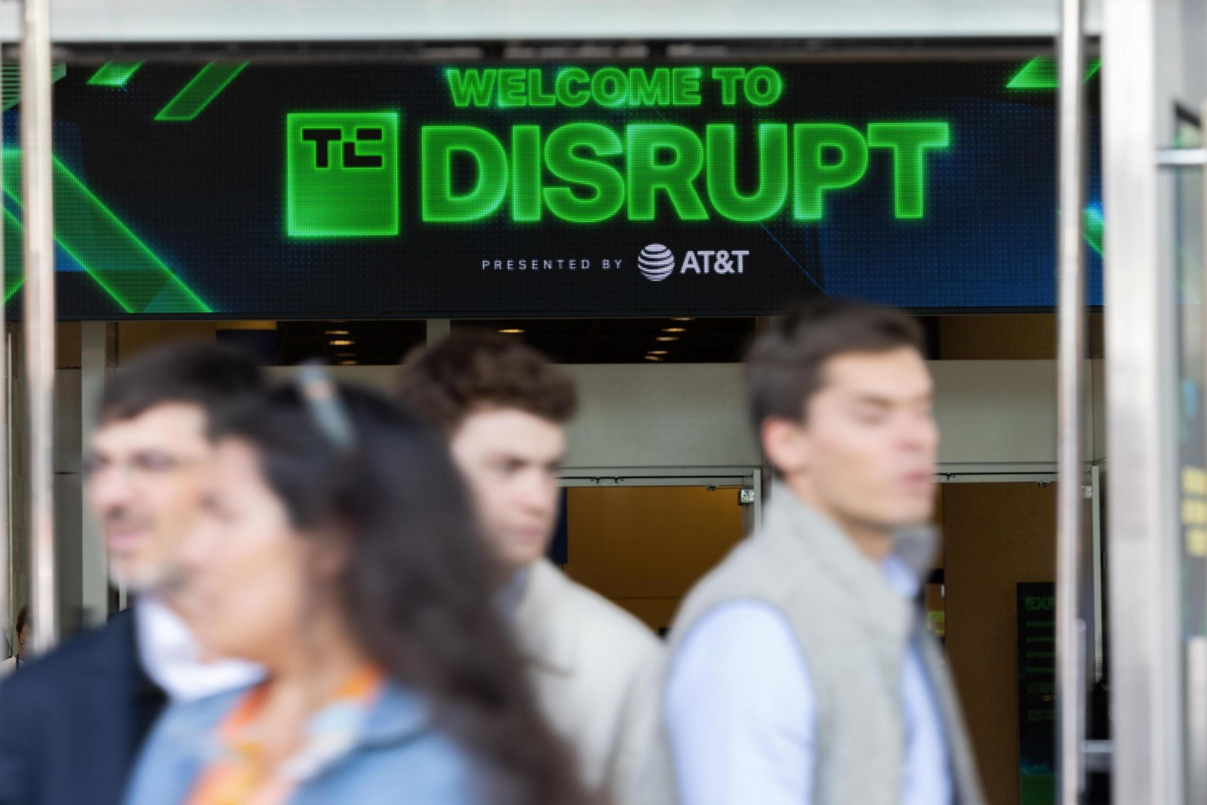 A digital sign reads "Welcome to TC Disrupt presented by AT&T" above an entrance, with four blurred people walking in front.