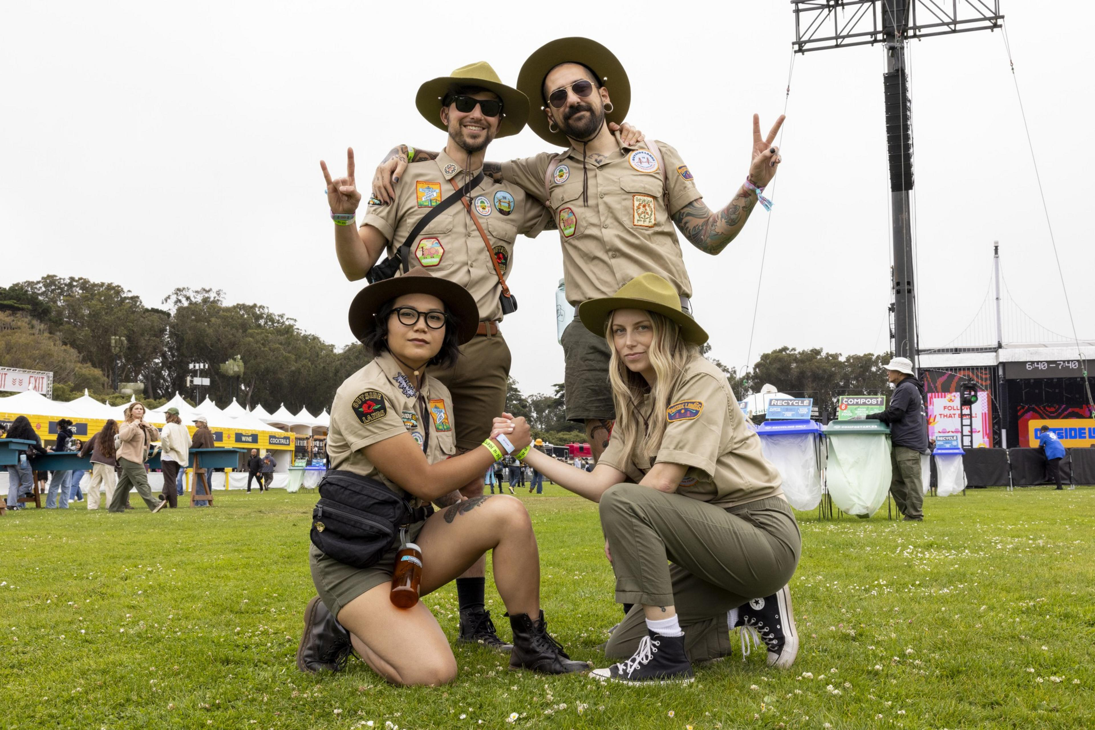 Four people in scout uniforms, two standing and two kneeling, pose on a grassy field at what appears to be an outdoor event with tents and a stage in the background.