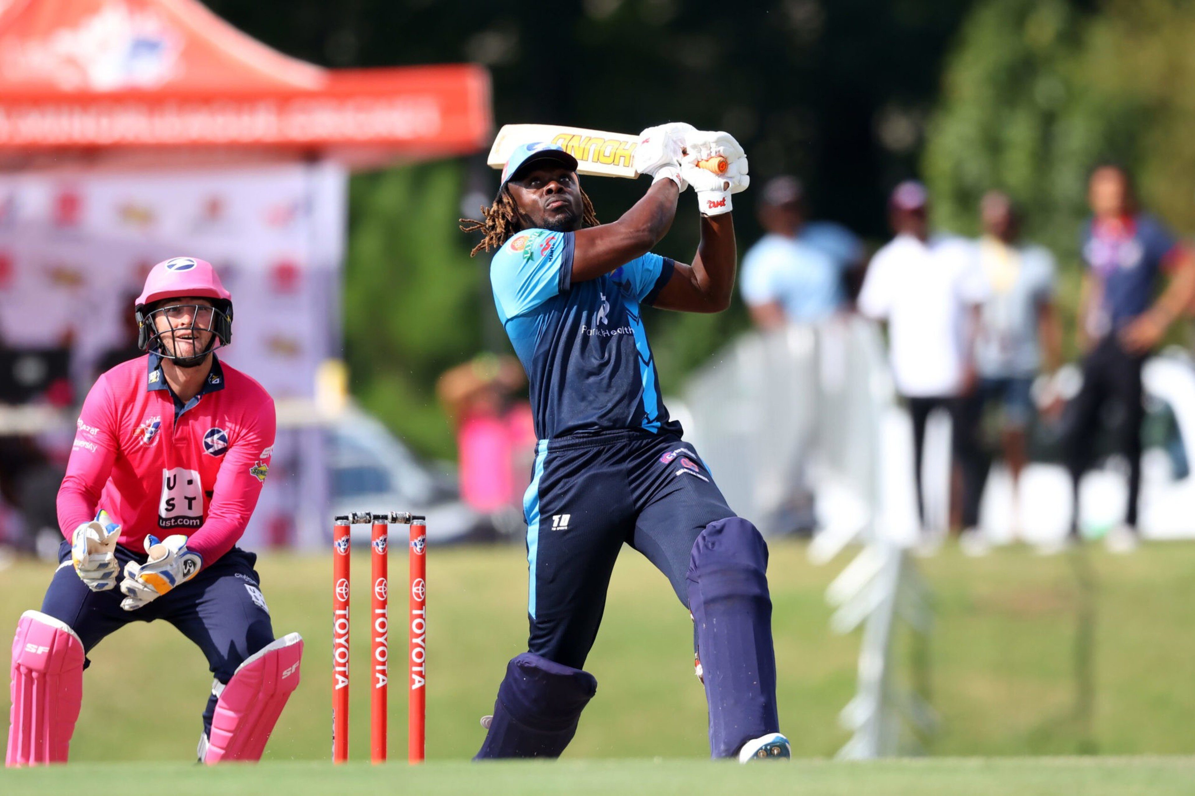 A cricket batsman in blue hits the ball while a wicketkeeper in pink crouches behind the stumps, with blurred spectators in the background.