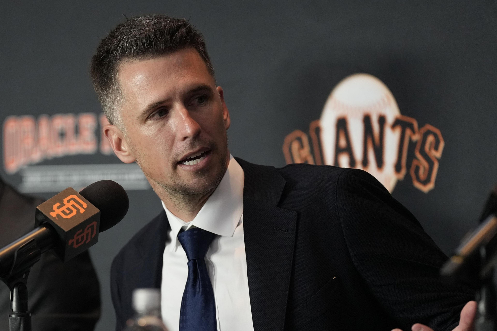 A man in a suit sits at a table with microphones, speaking at a press event. Behind him is a backdrop with the word "Giants" and a baseball graphic.