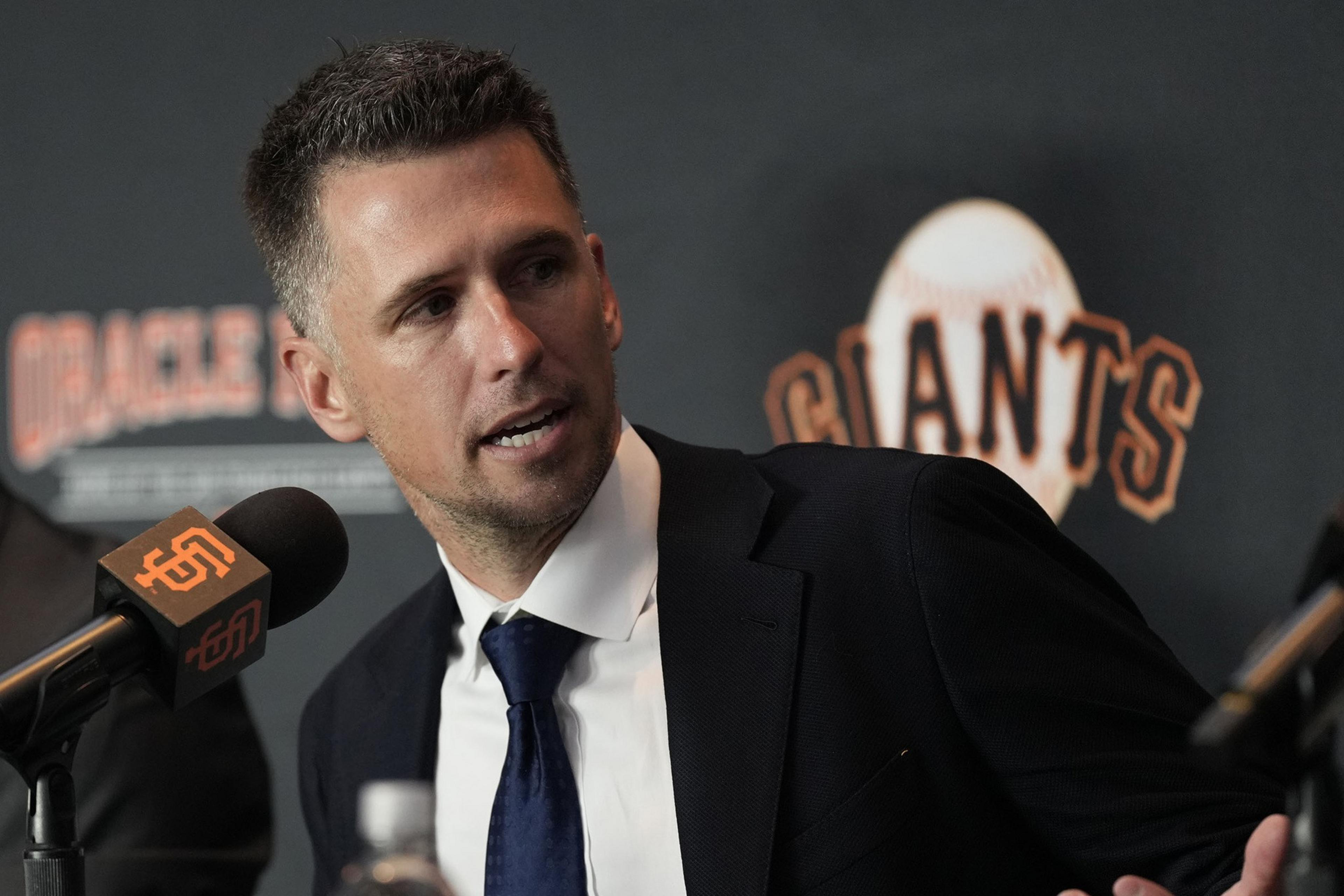 A man in a suit sits at a table with microphones, speaking at a press event. Behind him is a backdrop with the word "Giants" and a baseball graphic.