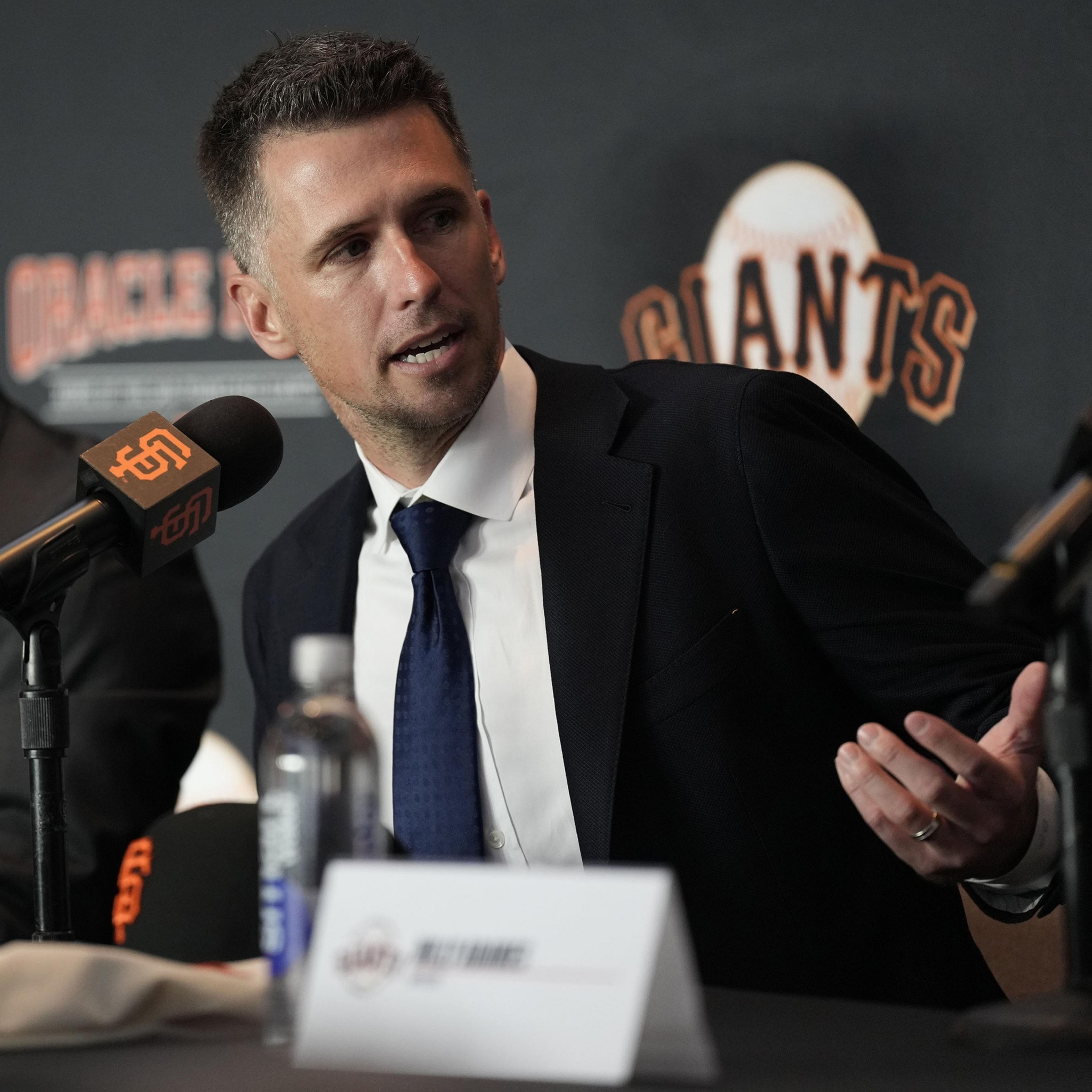 A man in a suit sits at a table with microphones, speaking at a press event. Behind him is a backdrop with the word "Giants" and a baseball graphic.