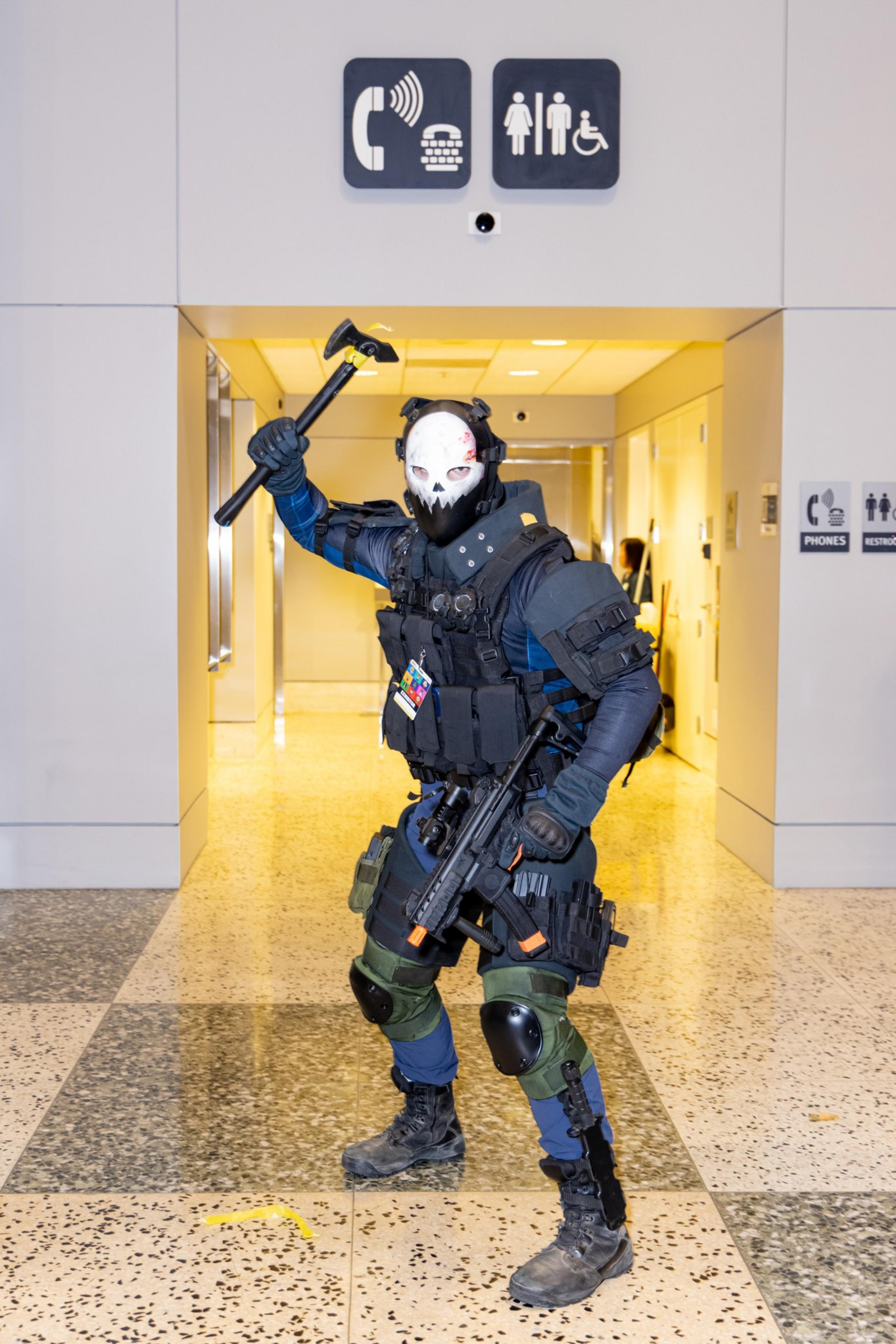 A person in tactical gear and a skull mask holds a tool resembling a small axe. They're standing in a hallway beneath signs for phones and restrooms.