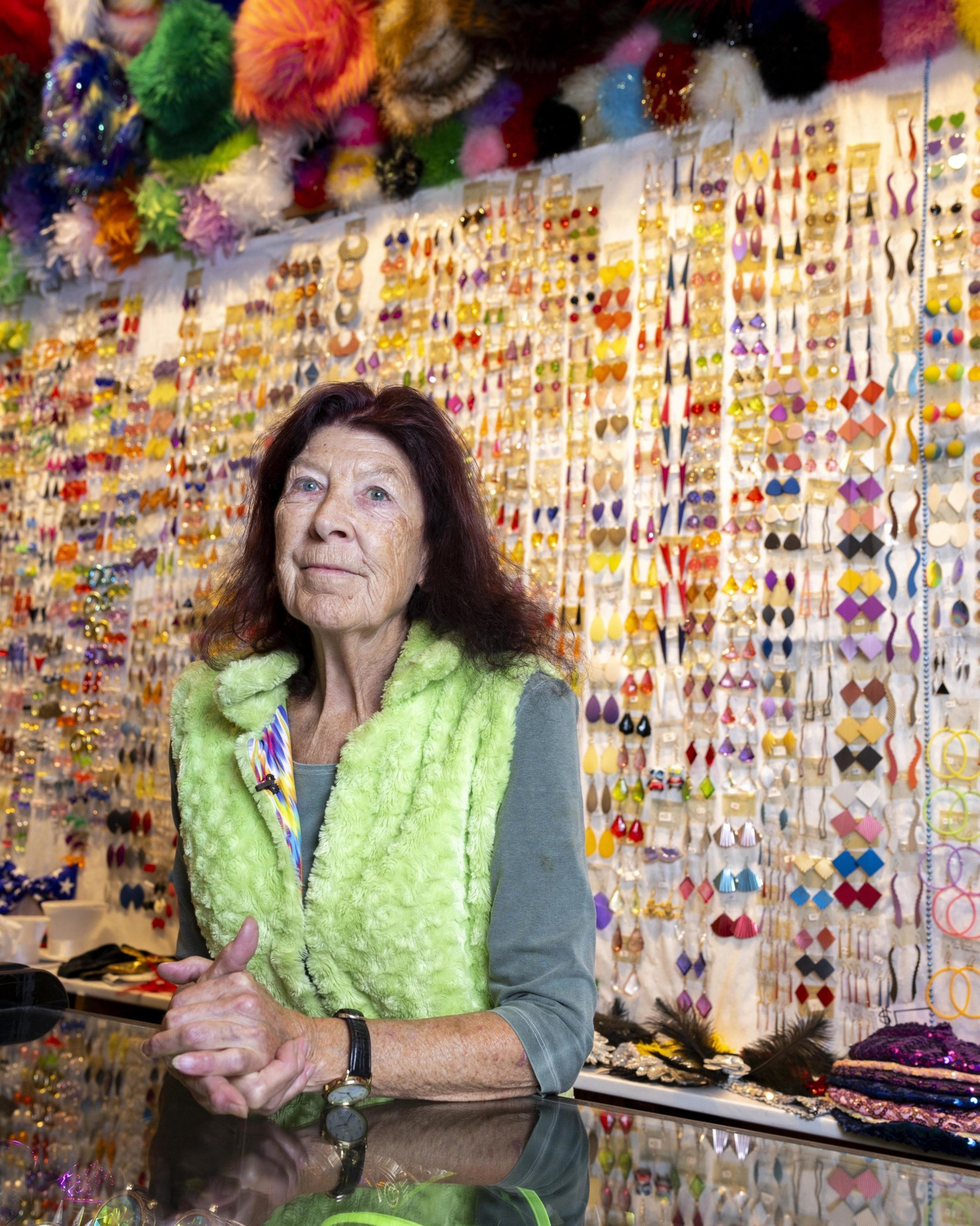 An elderly woman is standing behind a counter in front of a wall filled with colorful hanging earrings. She wears a lime green fuzzy vest and has variegated items above and around her.