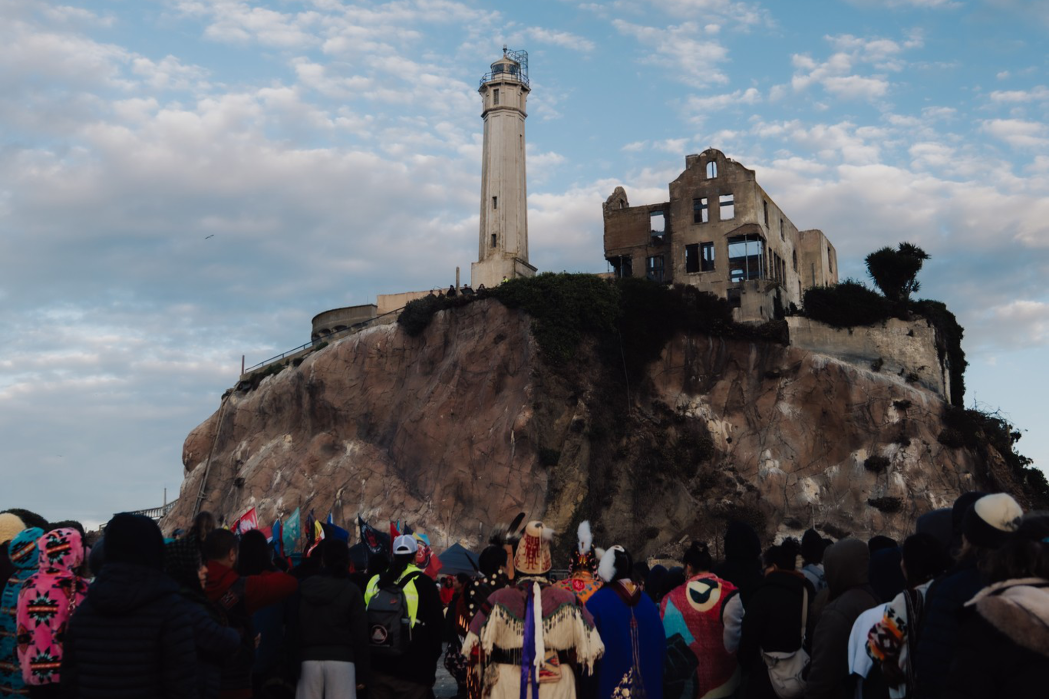A crowd of people faces a rocky island with a tall lighthouse and a partially ruined building under a sky filled with scattered clouds.