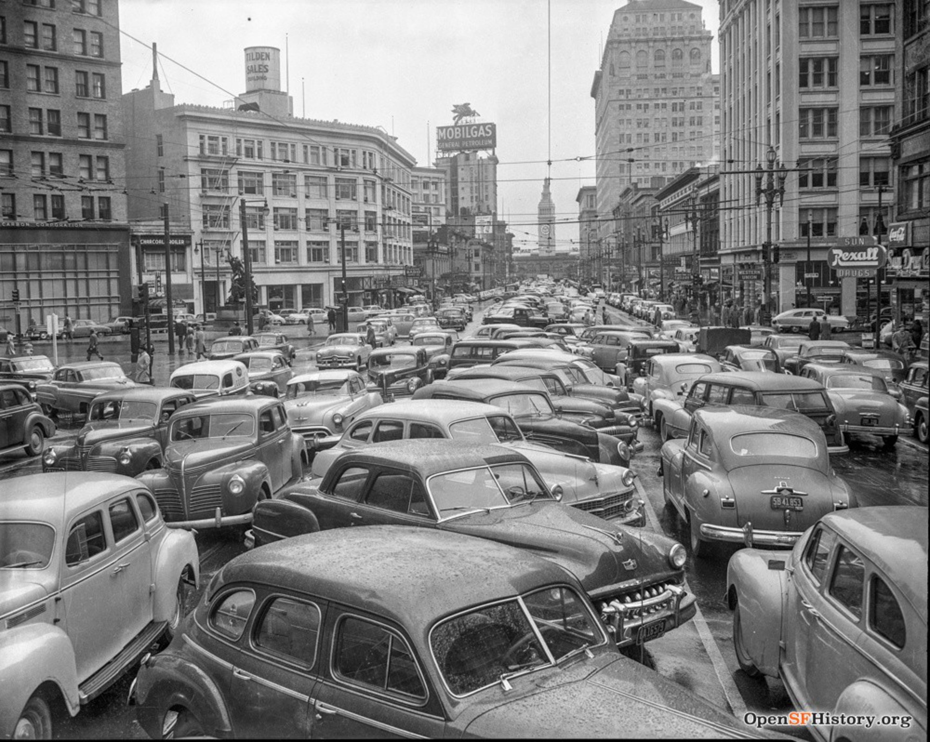 A crowded city street filled with tightly packed vintage cars and surrounded by tall buildings, with signs for Mobilgas and Rexall Drugs visible.