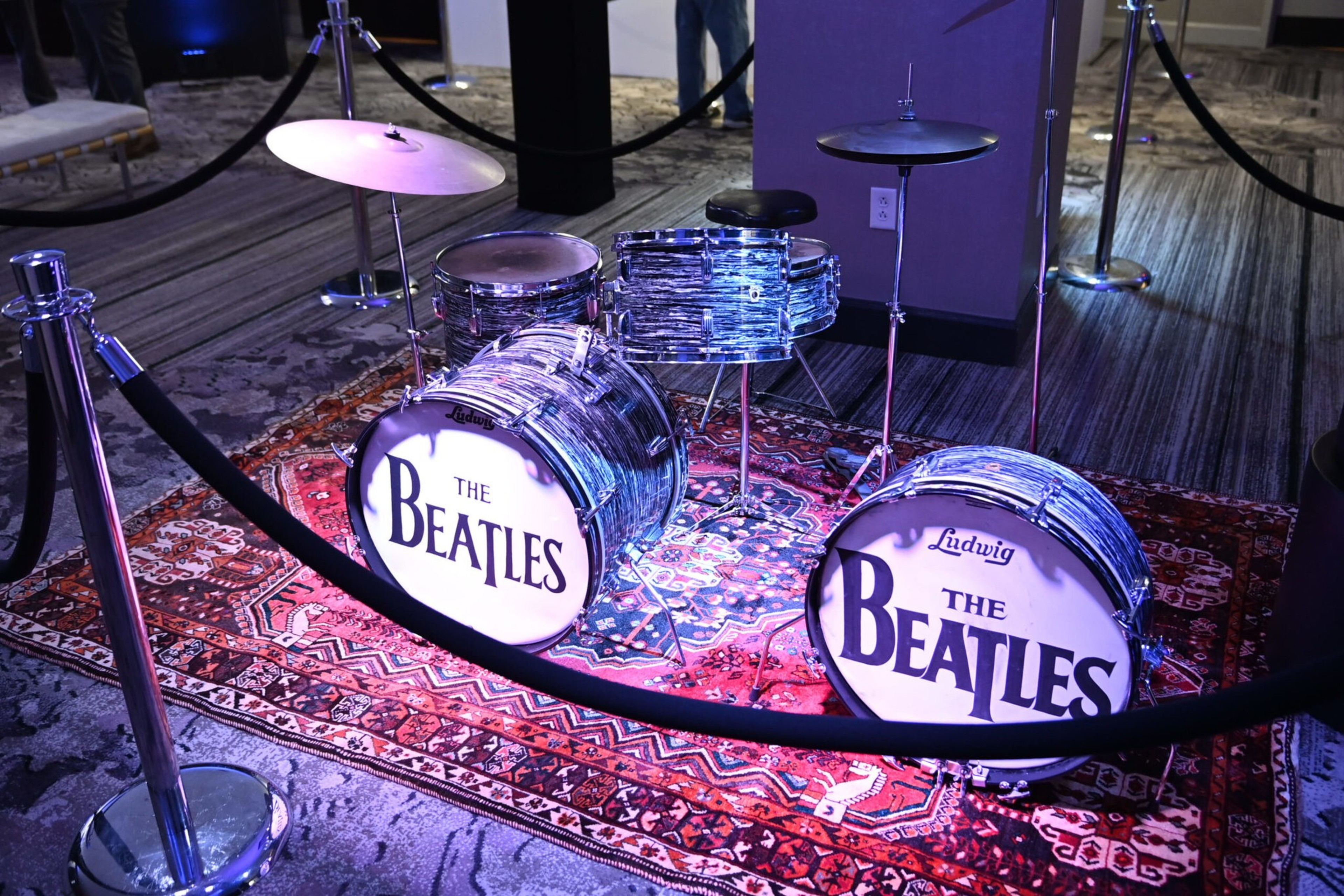 A vintage Ludwig drum set with "The Beatles" logo on the bass drums sits on a red patterned rug, surrounded by velvet ropes and a carpeted floor.