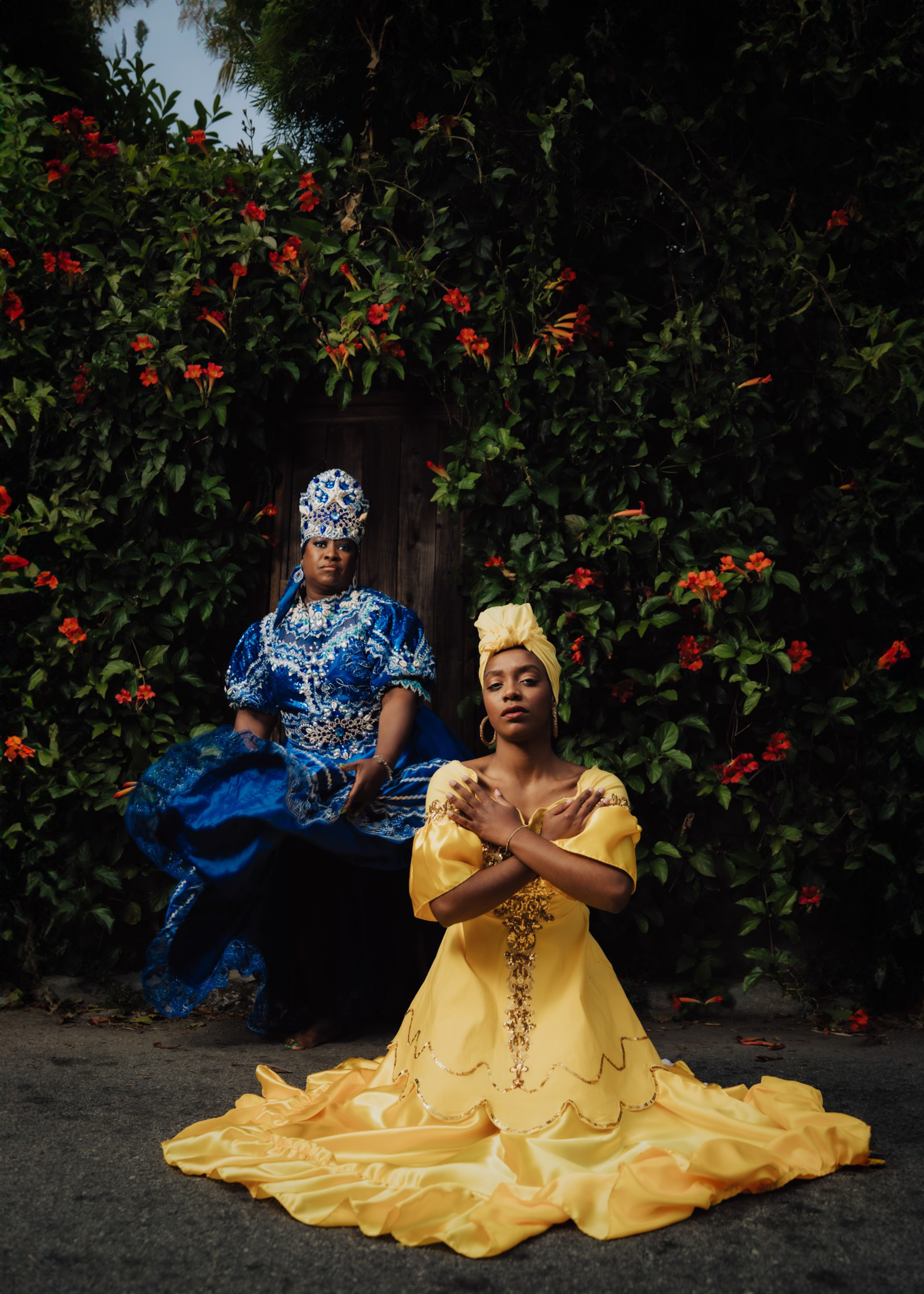 A woman in an elaborate yellow dress and headwrap kneels with crossed arms, while another woman in a detailed blue dress and headdress sits behind her.