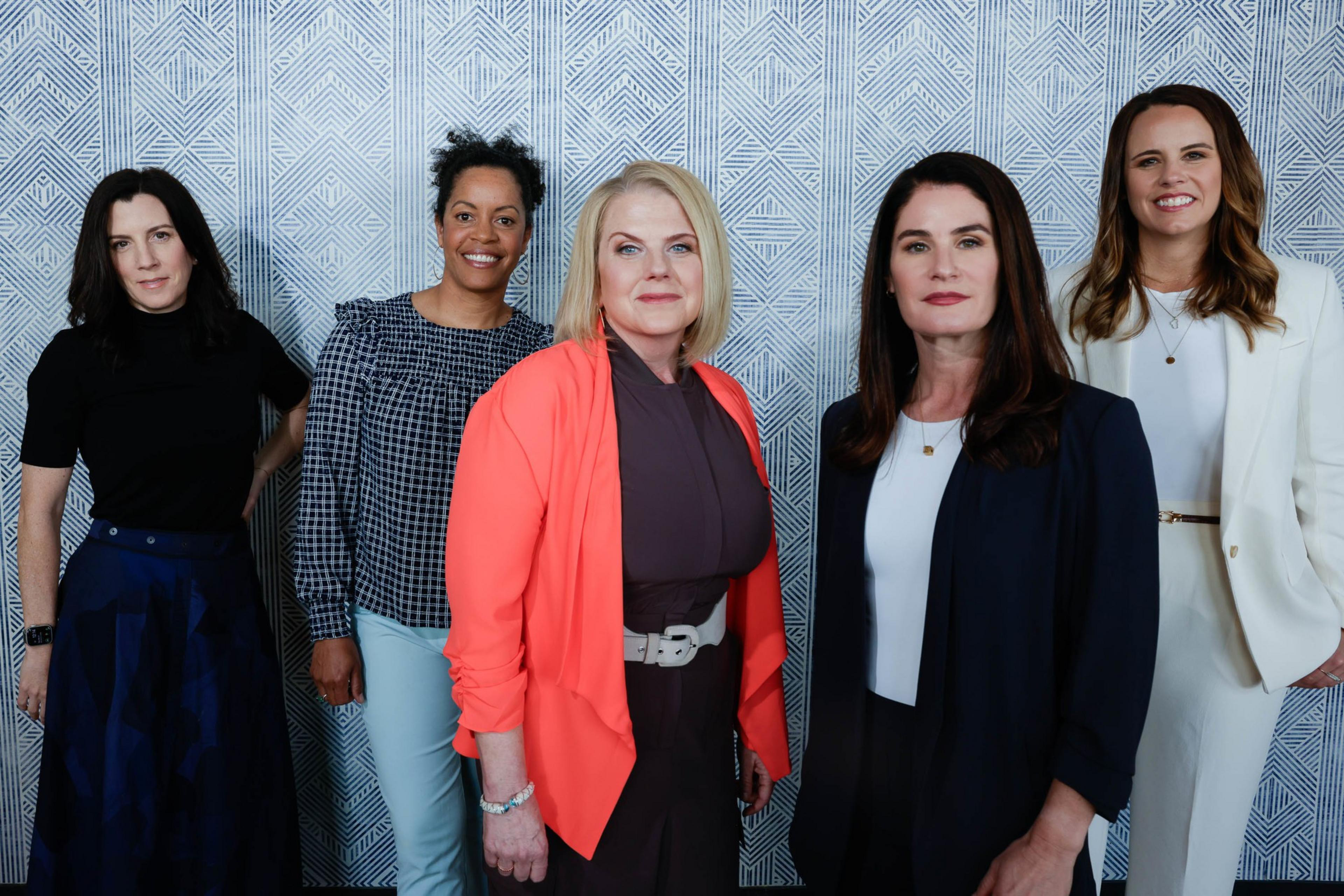 Five women stand smiling against a patterned backdrop, dressed in professional attire.