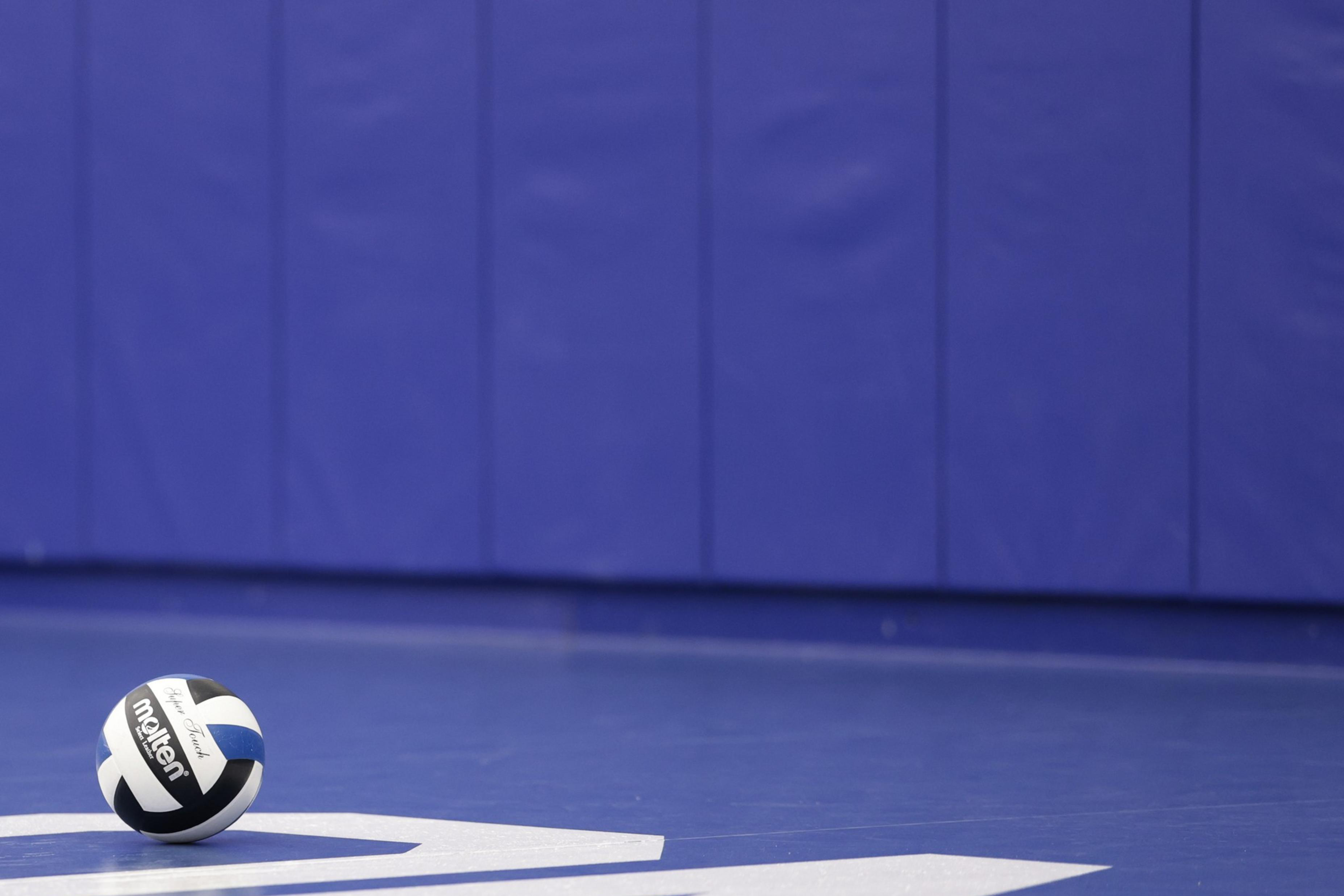 A volleyball rests on a blue gym floor with a padded blue wall in the background. The ball is black, white, and blue with the brand name visible.