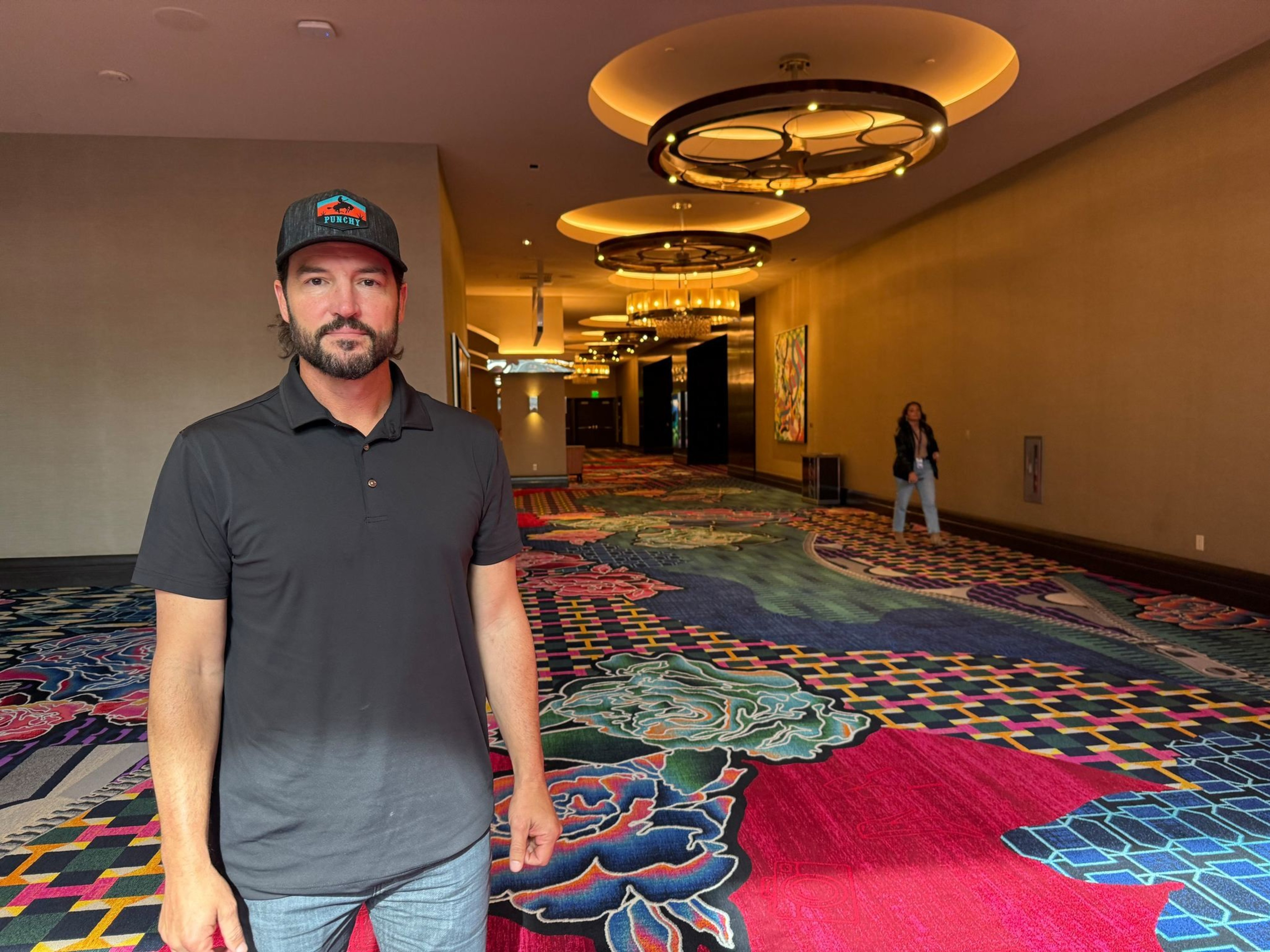 A man wearing a black polo and a cap stands on a colorful patterned carpet in a hallway with circular ceiling lights and a woman in the background.