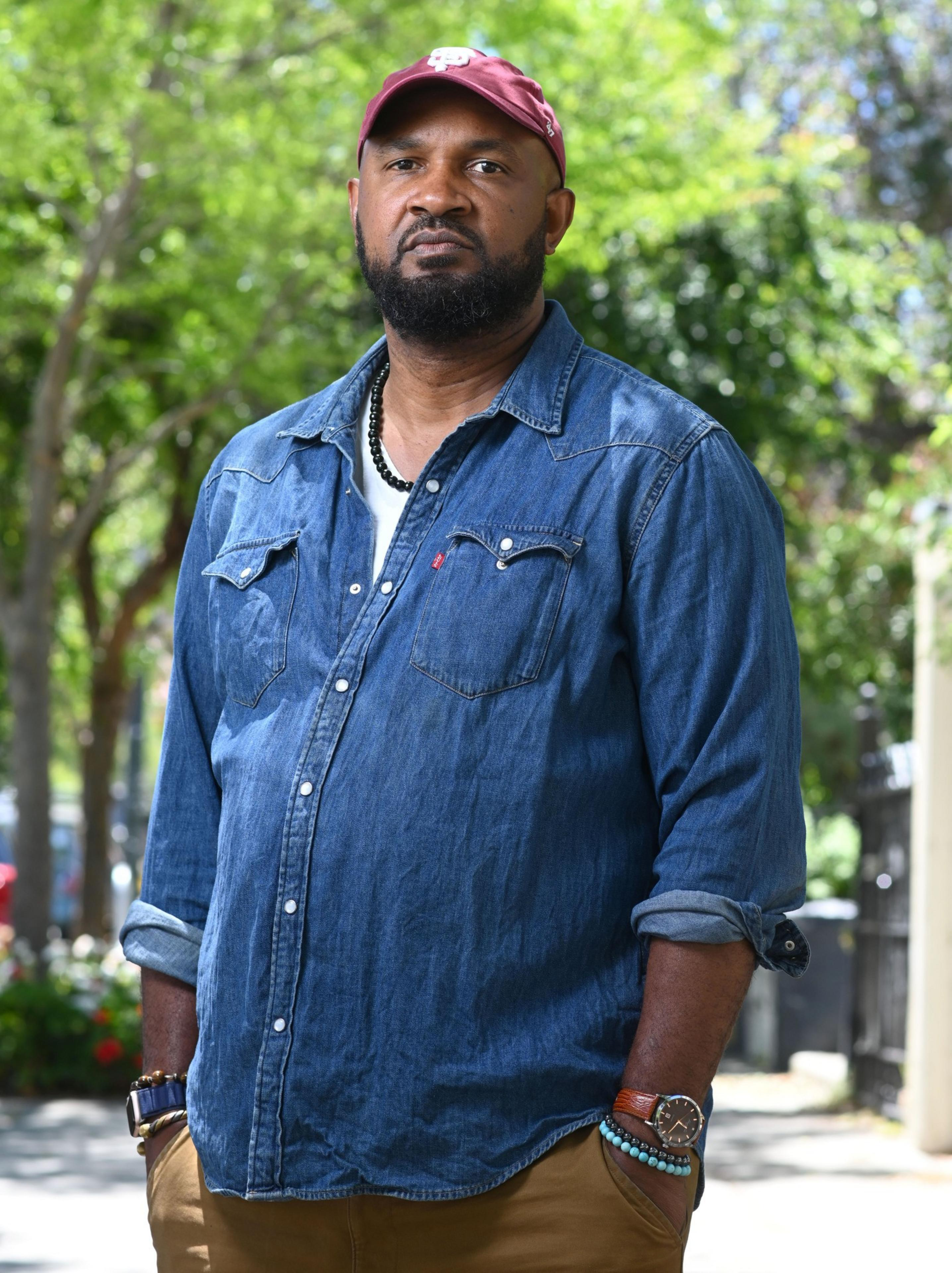 A man stands outdoors wearing a red cap, a blue denim shirt, brown pants, and beaded bracelets, with a backdrop of green trees and blurred foliage.