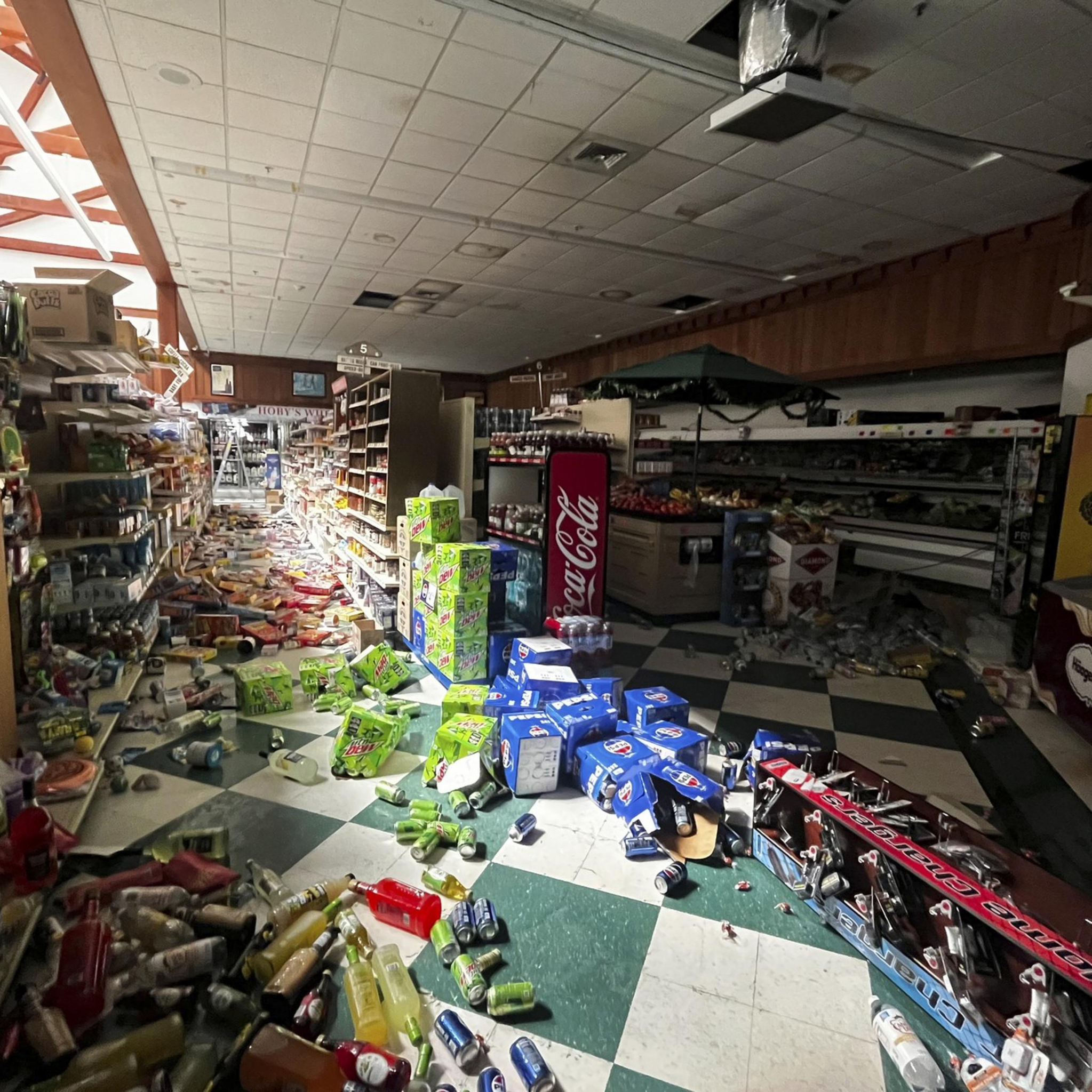 The image shows a store interior with products scattered across the floor, including knocked-over bottles and boxes. The ceiling appears damaged, and the scene is chaotic.