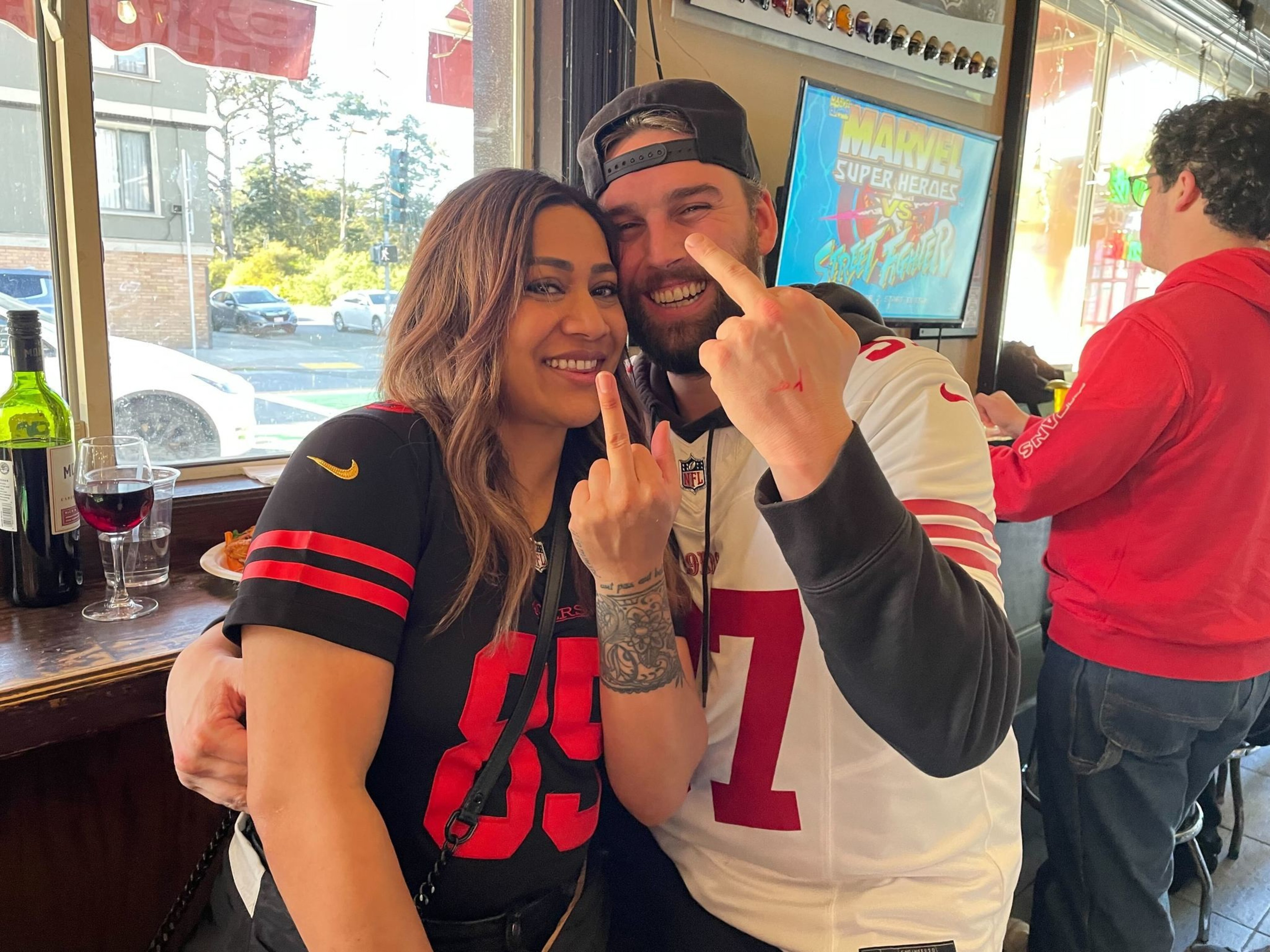 Two people are playfully gesturing at the camera, with sports jerseys on, inside a lively bar.