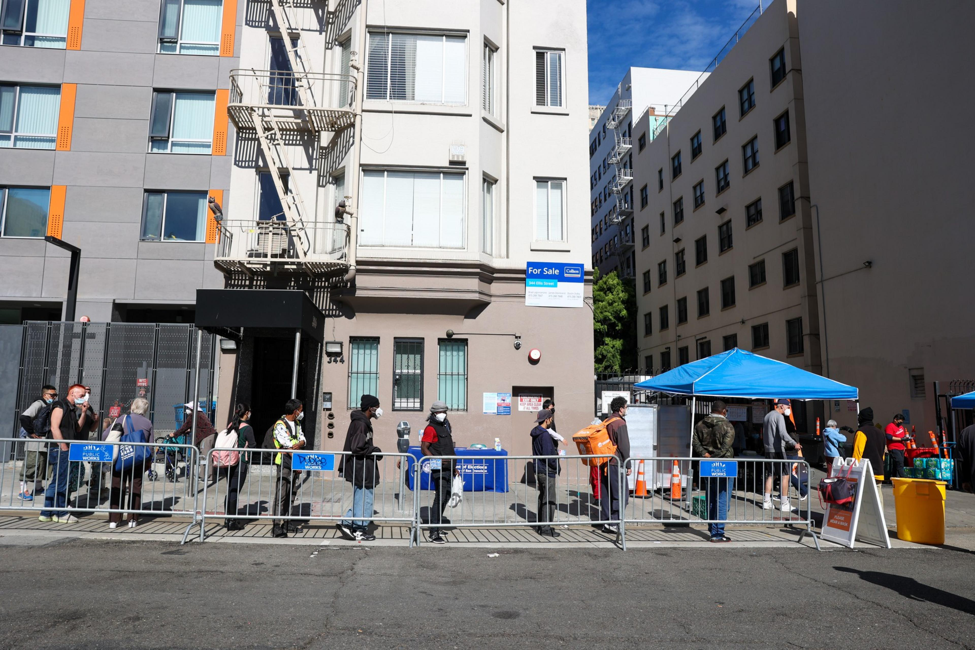 People wearing masks stand in a socially distanced line outside a building with a "For Sale" sign, next to a blue tent on a sunny day.
