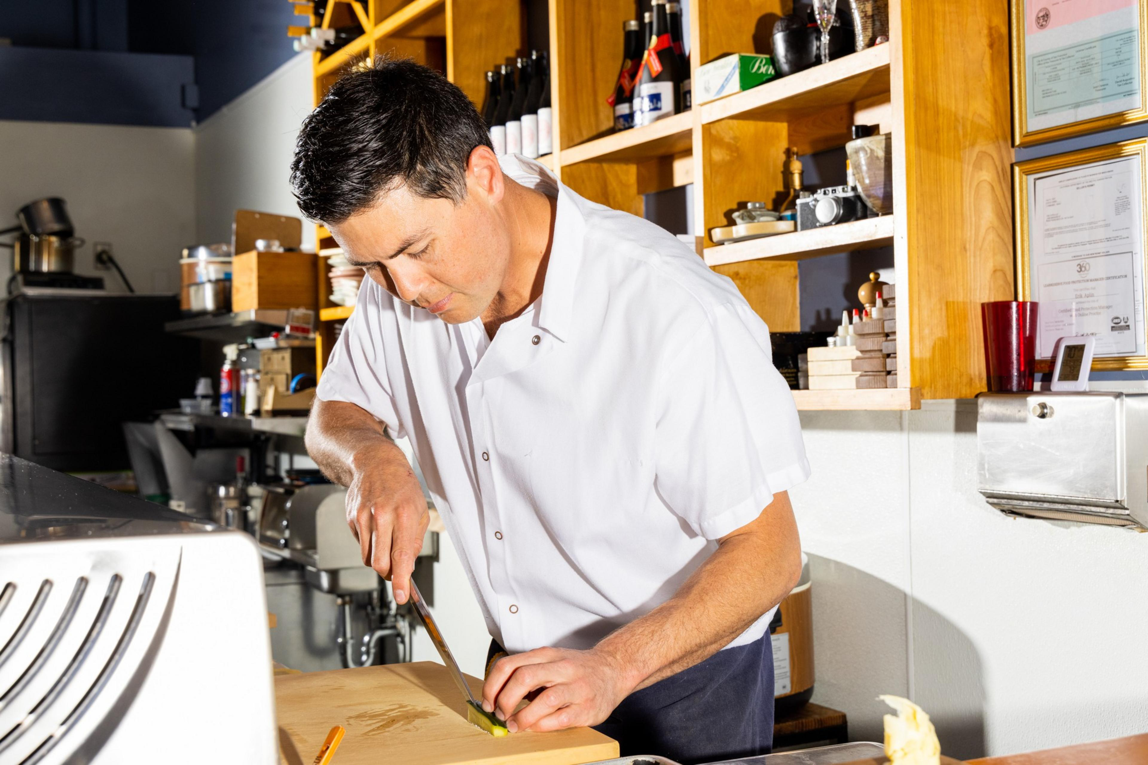 A chef in a white uniform is carefully slicing vegetables on a wooden cutting board in a well-organized kitchen with shelves of various items behind him.