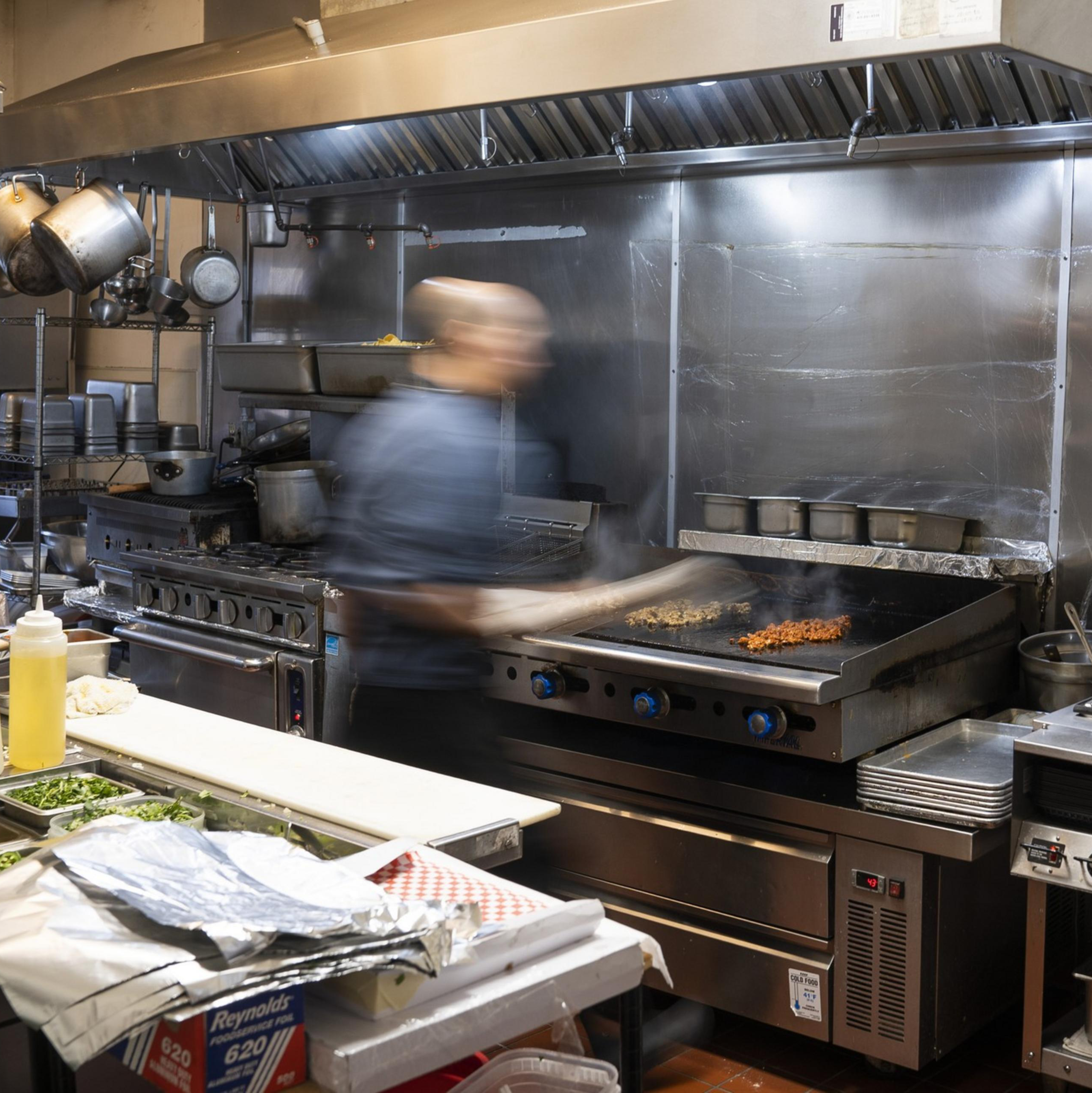 A chef in a bustling kitchen is cooking on a large grill, with lots of metal cookware around. Ingredients are prepared on the counter, and steam rises from the food.