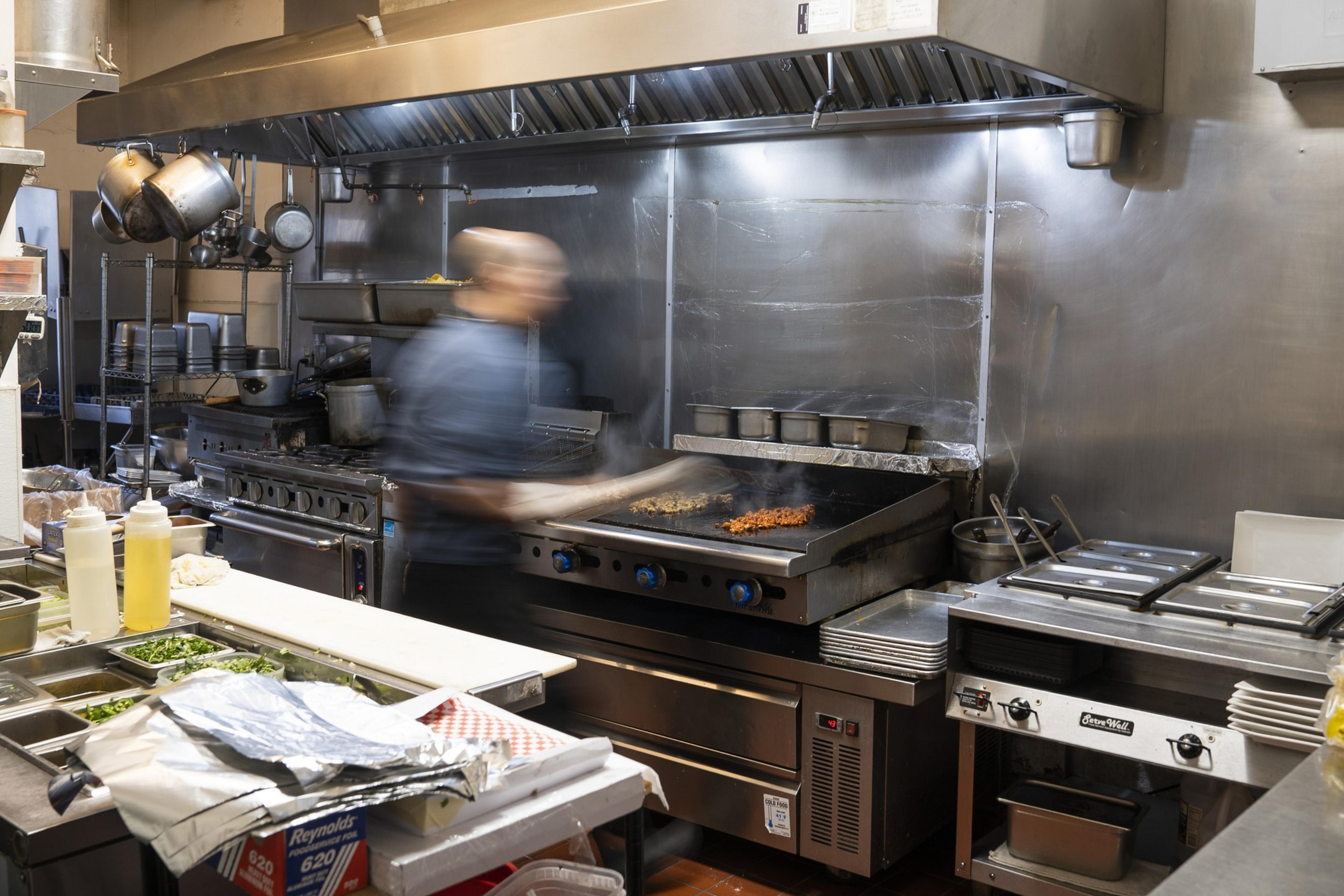 A chef in a bustling kitchen is cooking on a large grill, with lots of metal cookware around. Ingredients are prepared on the counter, and steam rises from the food.