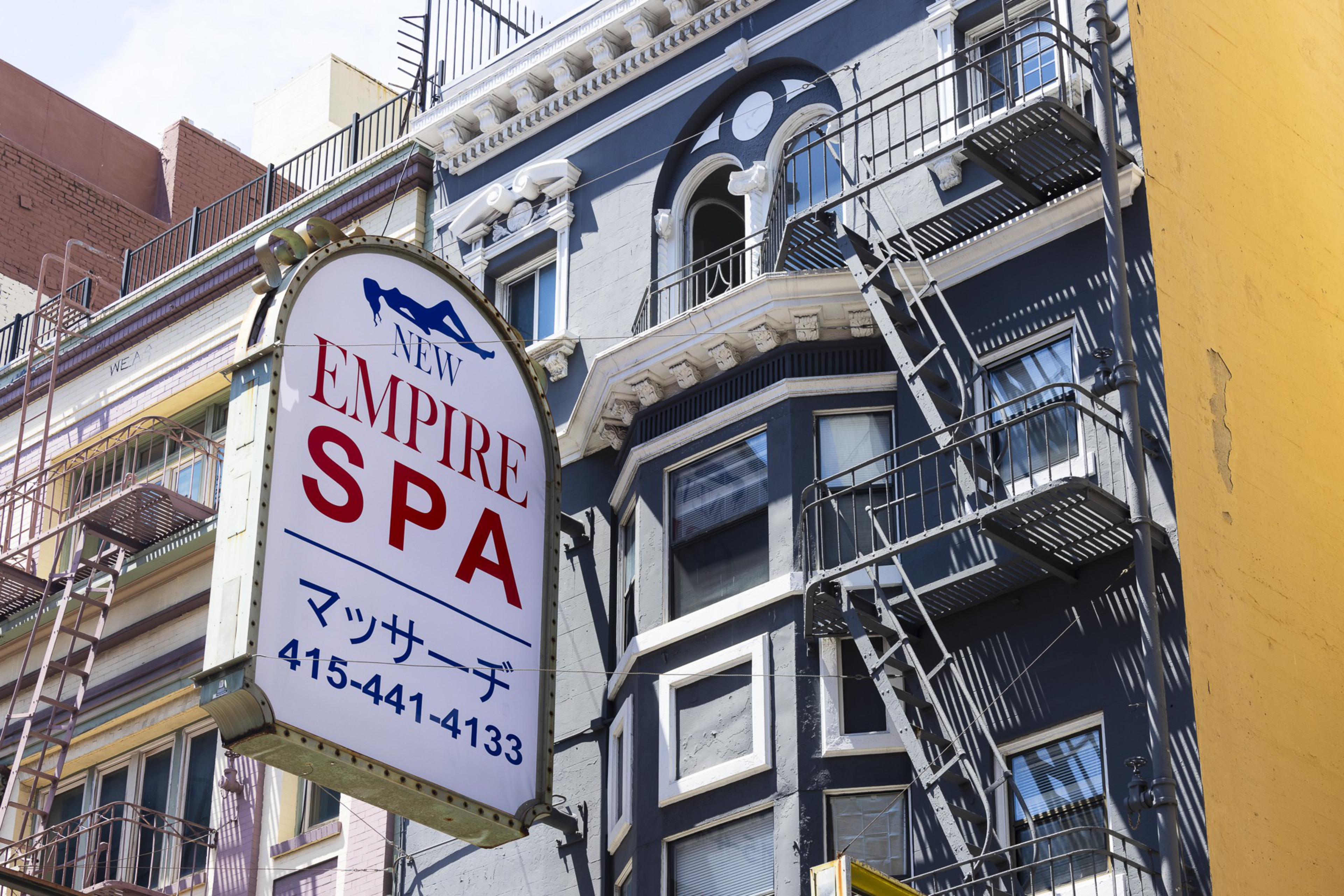 A sign for &quot;New Empire Spa&quot; is mounted on a colorful building with fire escapes, featuring Japanese text and a contact number.