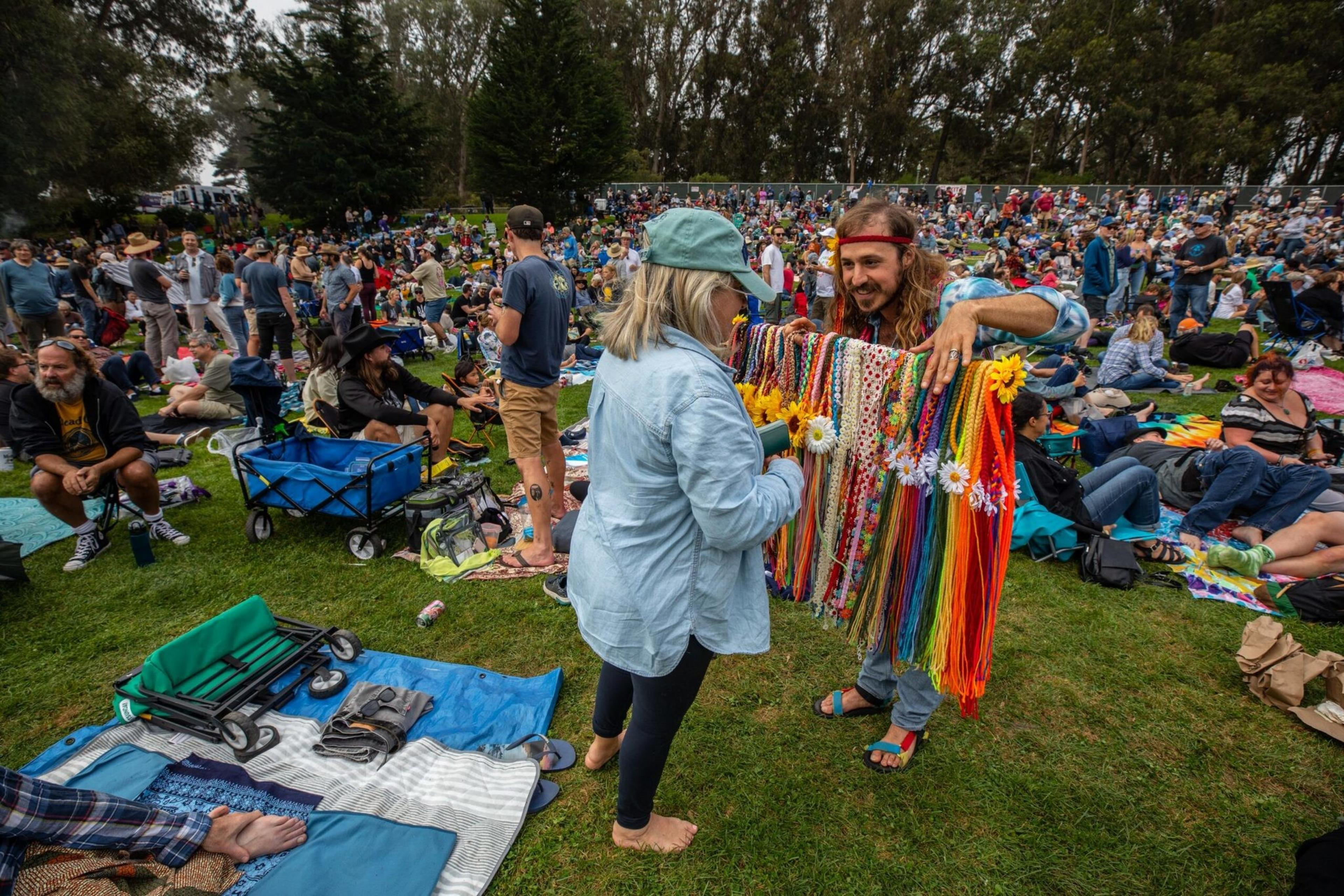 A man displays colorful, beaded necklaces decorated with flowers to a woman in a crowded outdoor grassy area with many people sitting on blankets.
