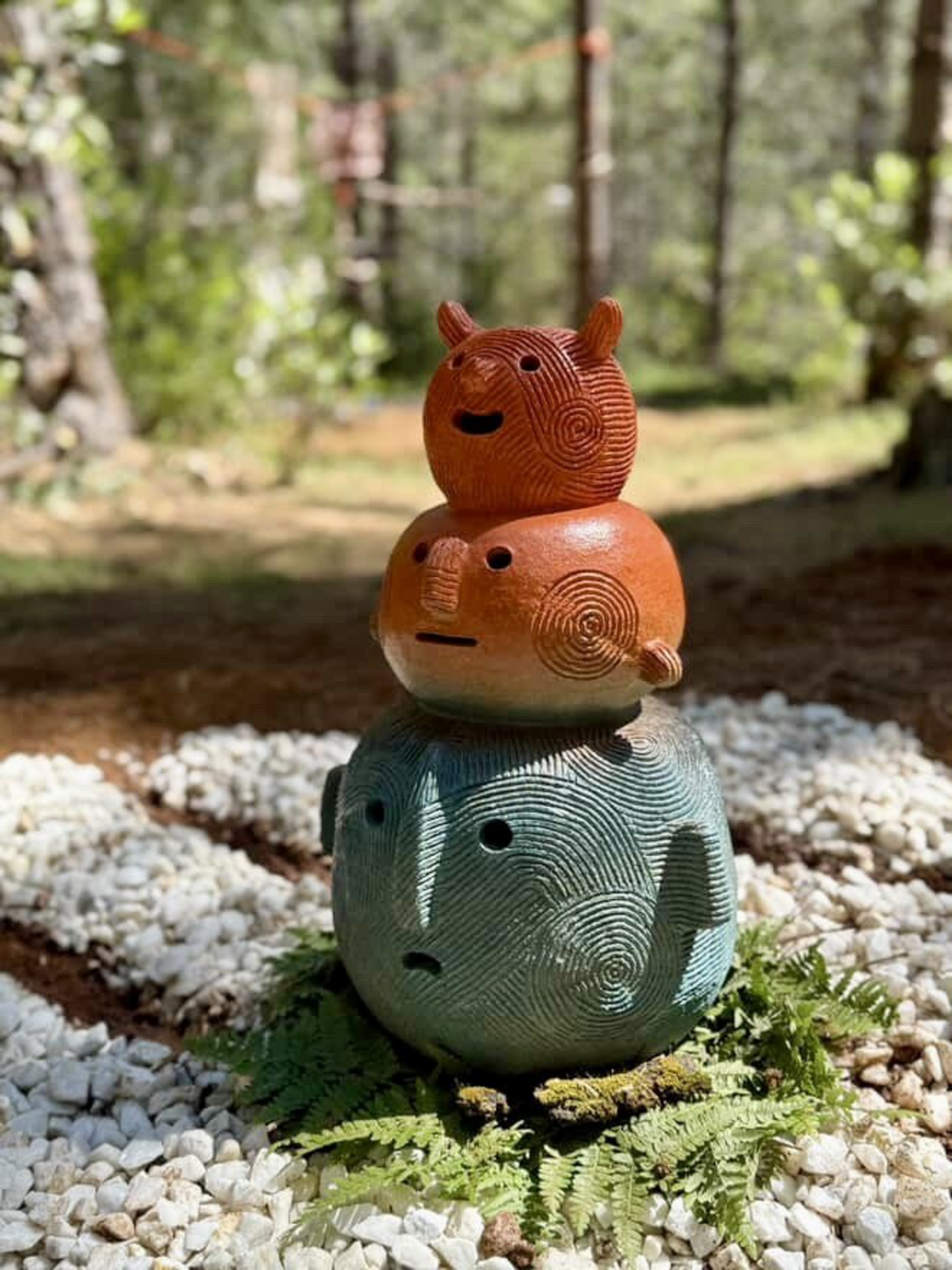 Three textured, rounded ceramic sculptures with faces and arms are stacked vertically on green ferns surrounded by white pebbles in a forest.