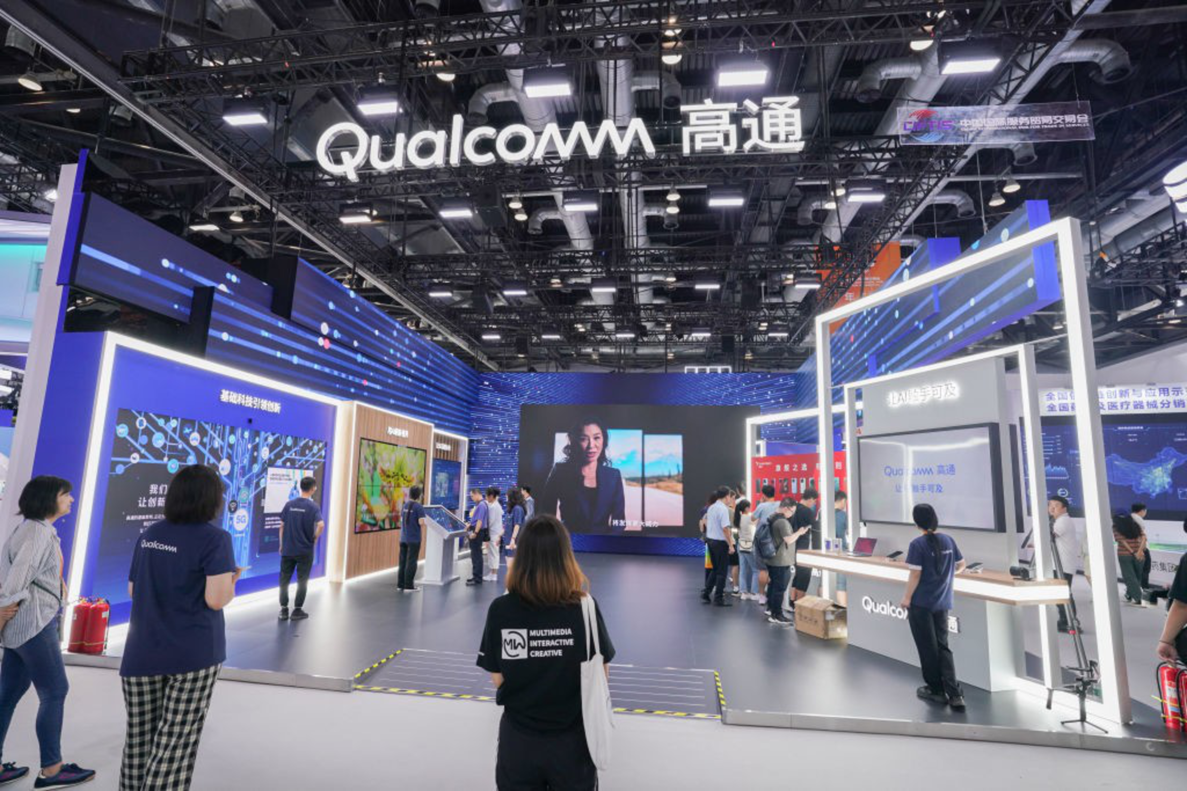 A modern Qualcomm booth with bright LED displays and visitors exploring various technology exhibits in a spacious, well-lit exhibition hall.