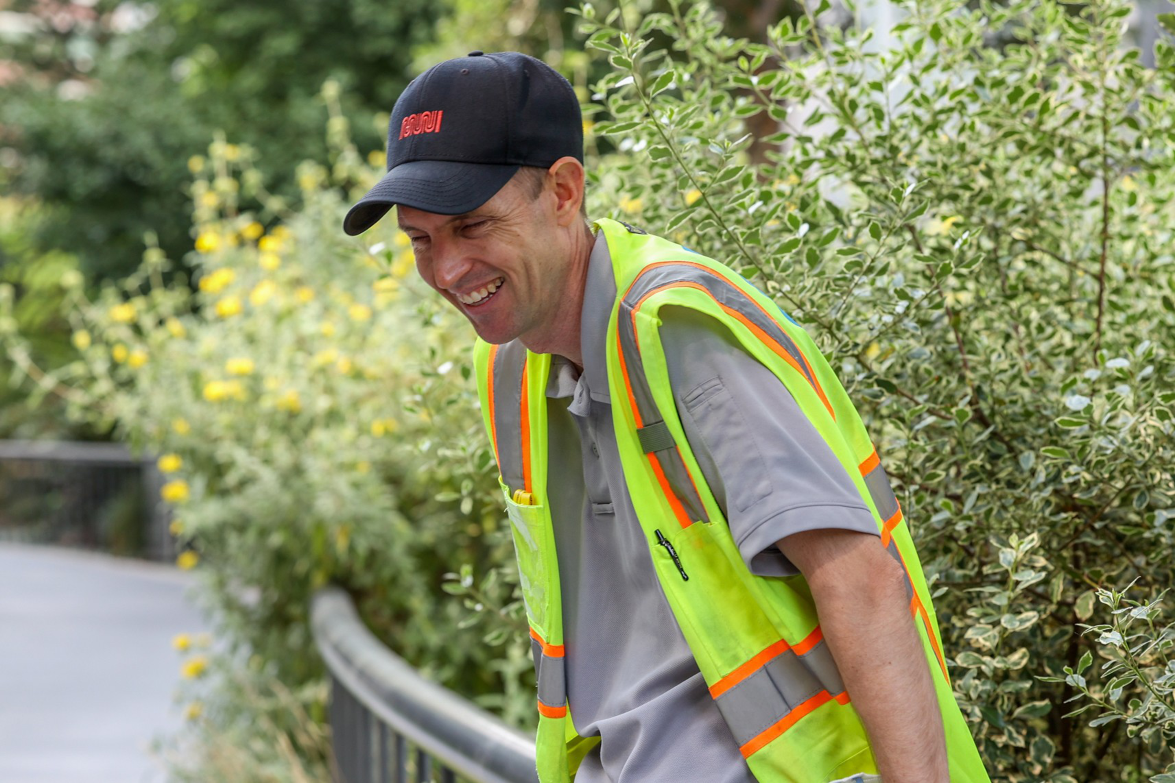 A man is smiling, wearing a black cap and a reflective safety vest over a gray shirt. He's standing outdoors near green bushes and yellow flowers.