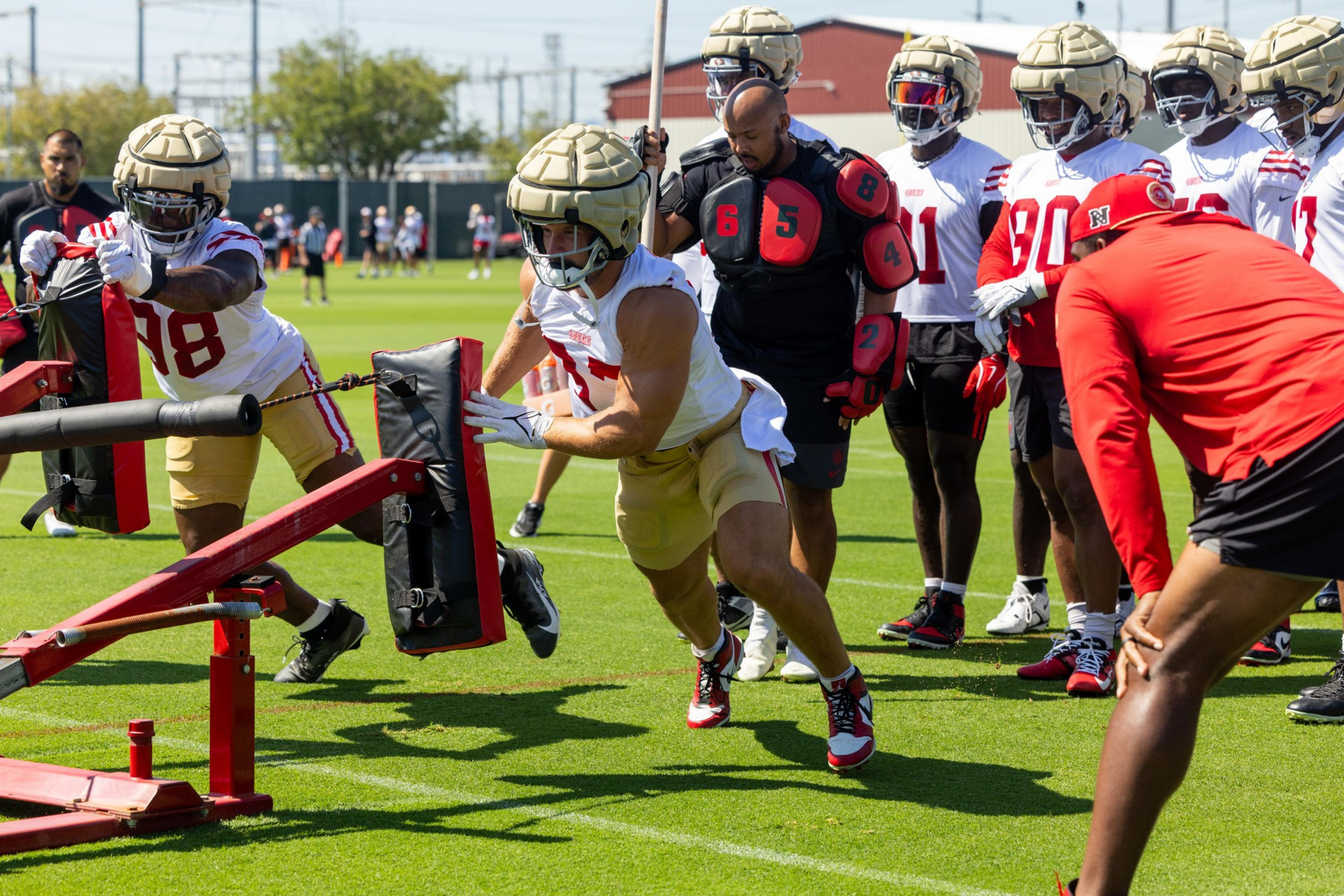 Football players are practicing on a grassy field, wearing helmets with pads. They are engaged in a drill, pushing against a blocking sled. Coaches observe.