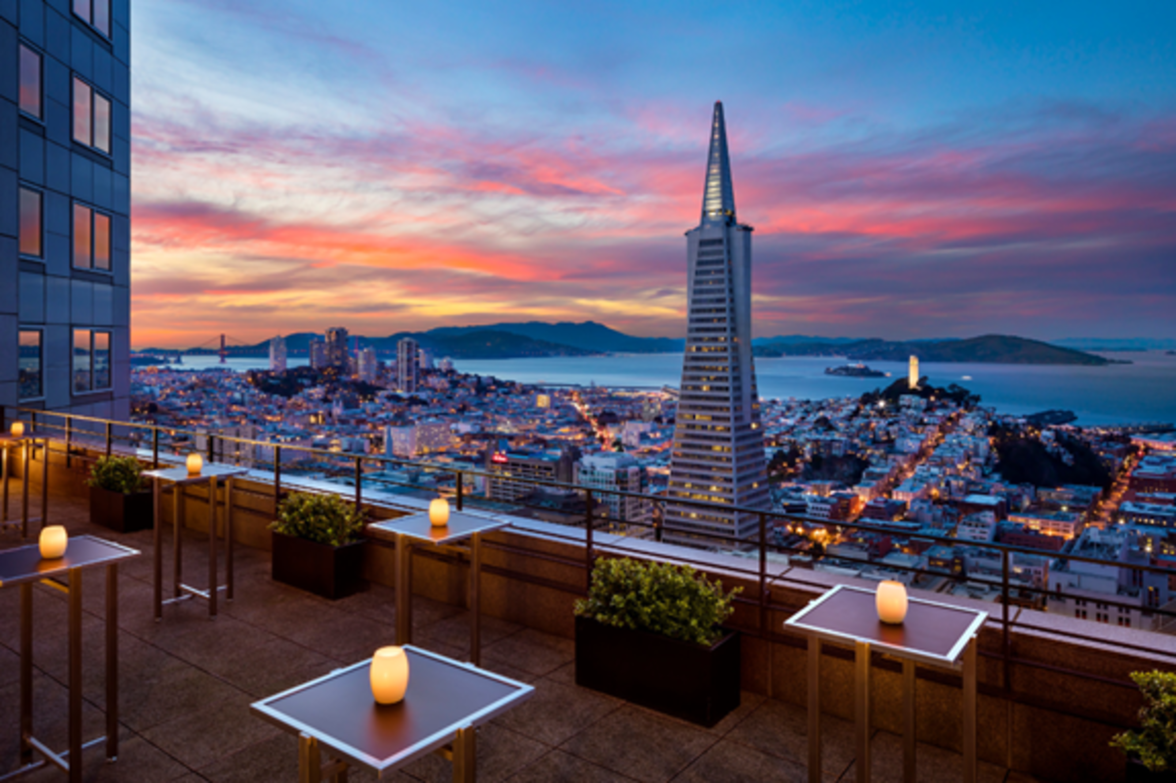 A rooftop terrace with small tables and glowing candles overlooks a cityscape featuring a tall, pointed skyscraper, a sunset sky, water, and distant hills.