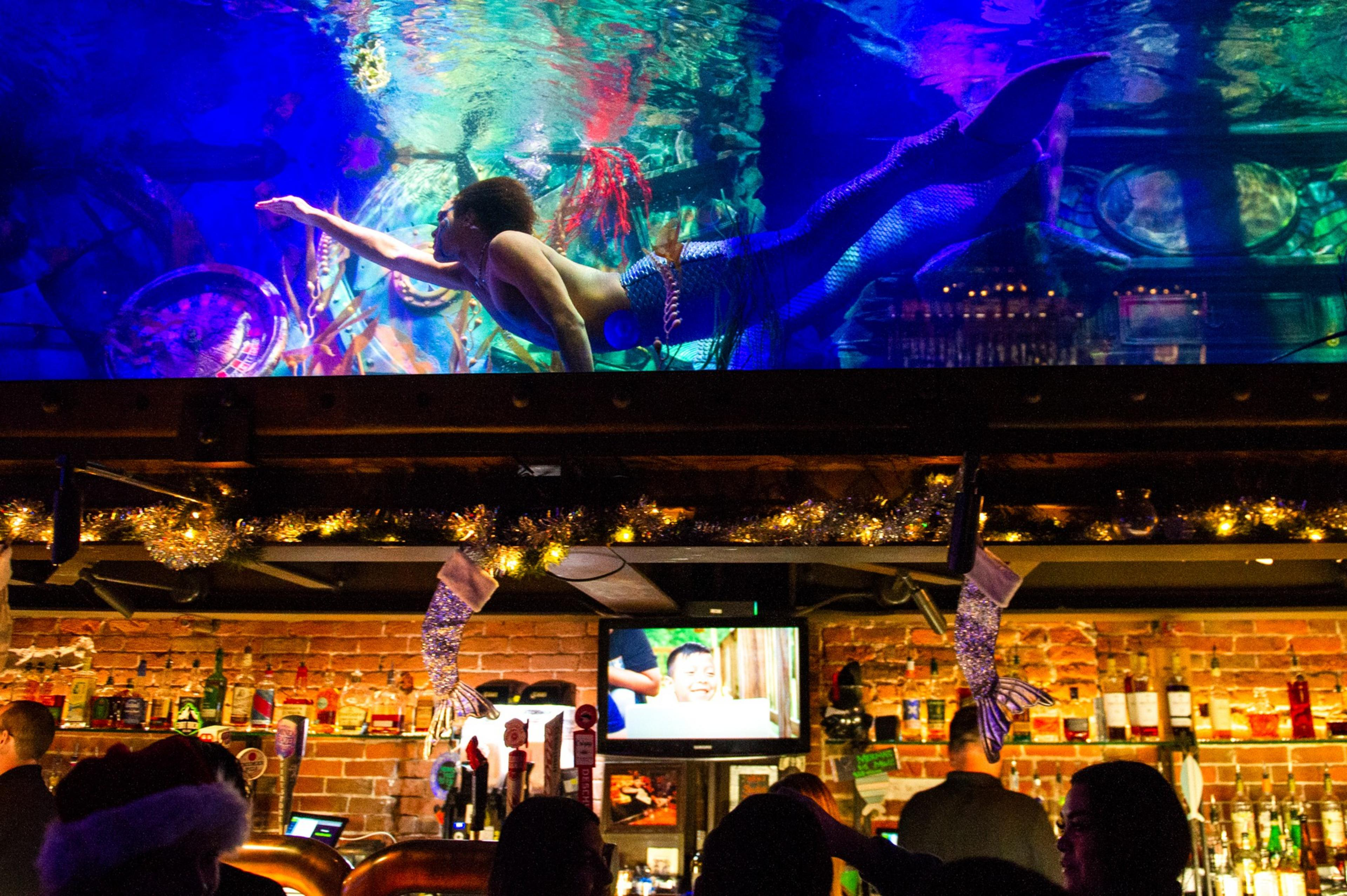 A mermaid swims in a brightly lit tank above a bar decorated with festive garlands and mermaid tail ornaments, with patrons below.
