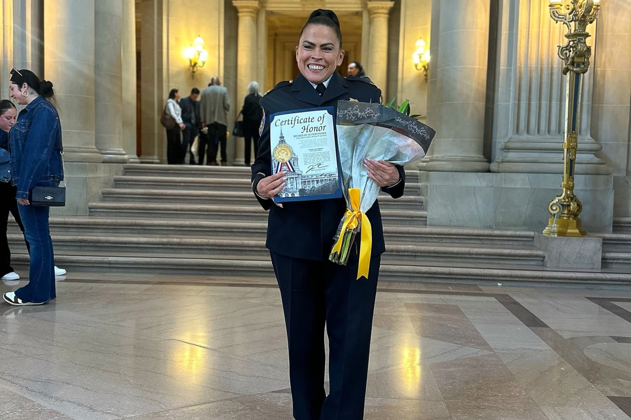 A smiling woman in a dark uniform holds a Certificate of Honor and a bouquet with a yellow ribbon, standing in a grand hall with columns and steps behind her.