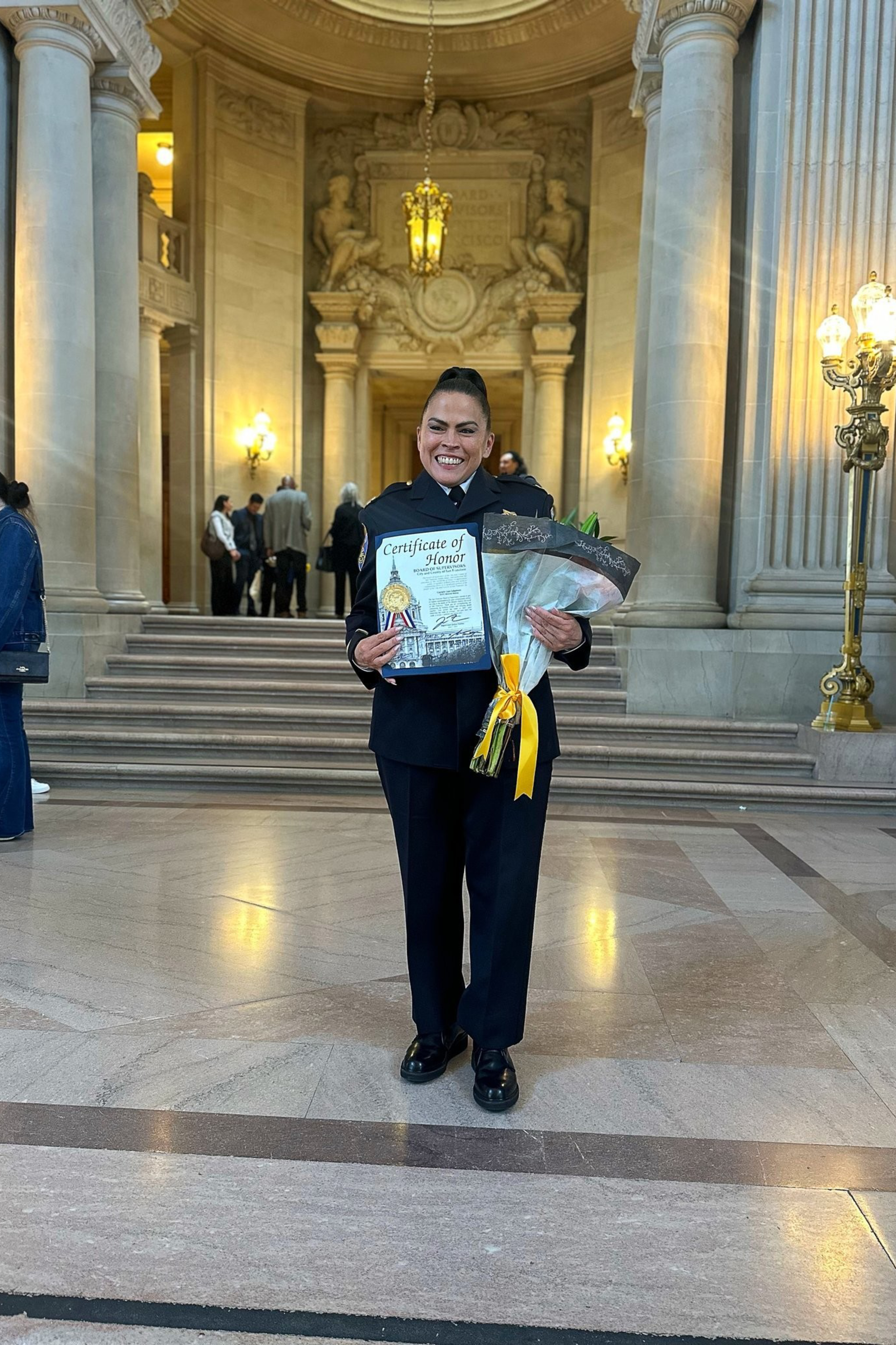 A smiling woman in a dark uniform holds a Certificate of Honor and a bouquet with a yellow ribbon, standing in a grand hall with columns and steps behind her.