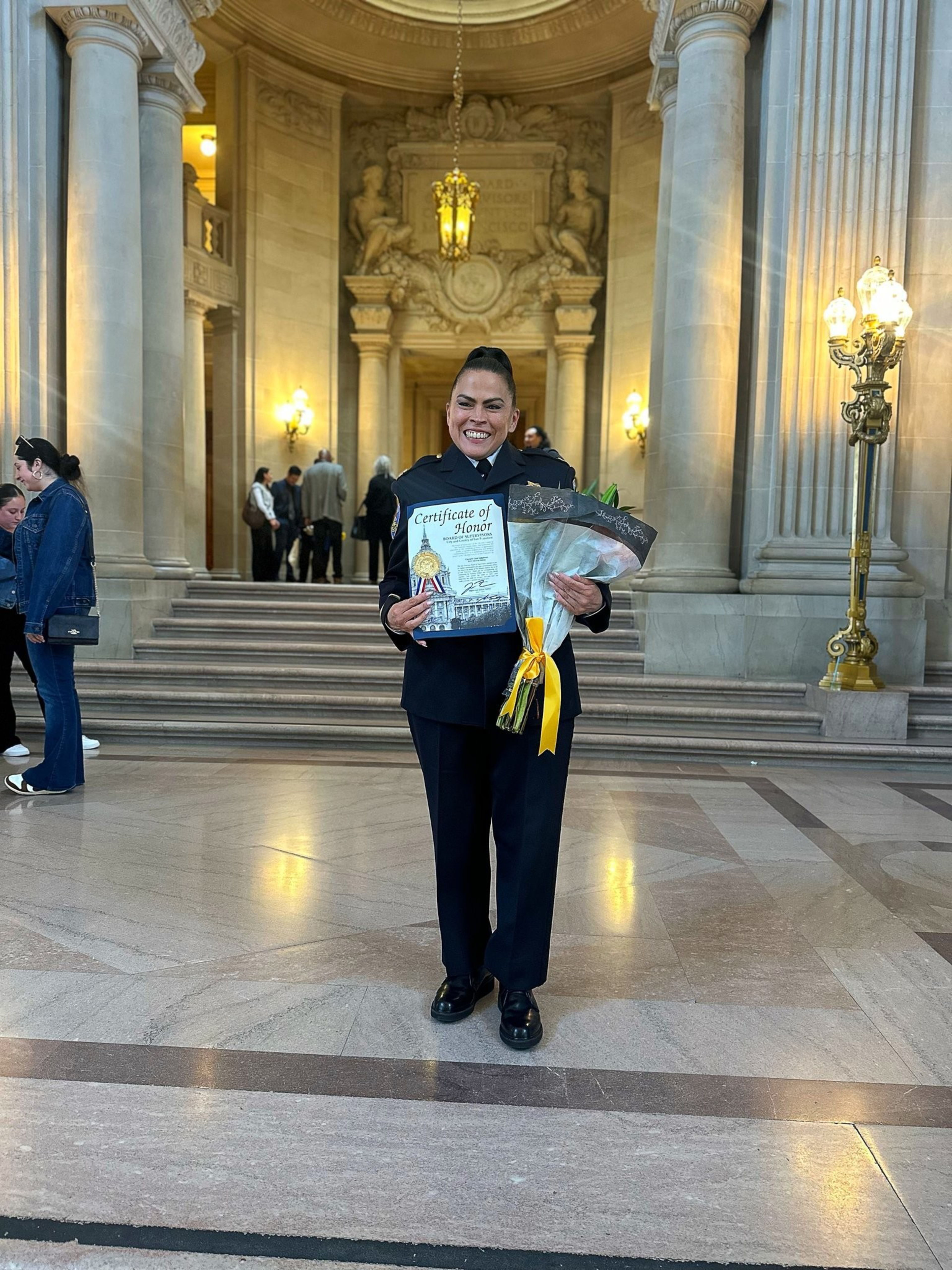 A smiling woman in a dark uniform holds a Certificate of Honor and a bouquet with a yellow ribbon, standing in a grand hall with columns and steps behind her.