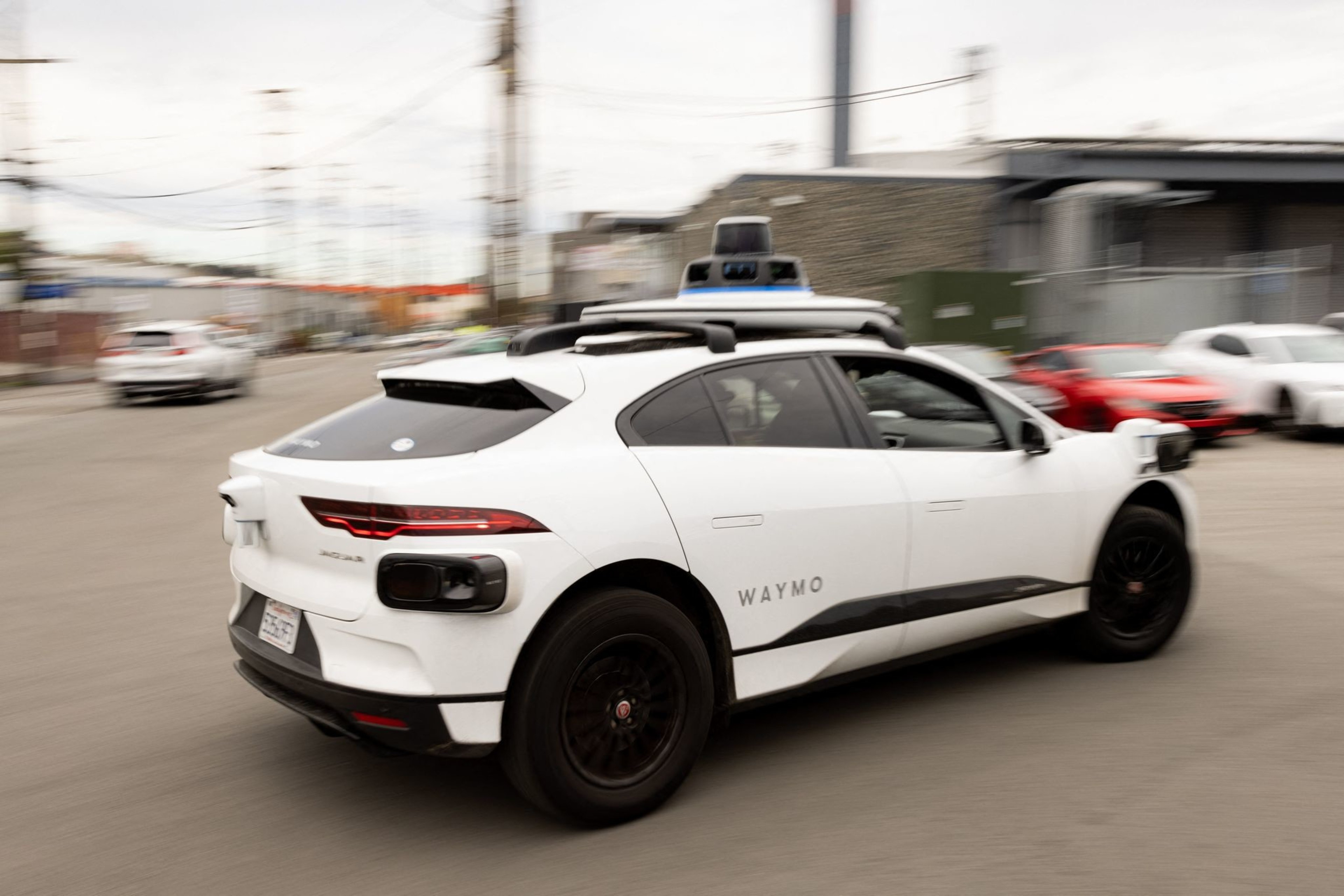 A white Waymo-branded self-driving car with sensors on the roof is driving on a street with several parked cars and industrial buildings in the background.
