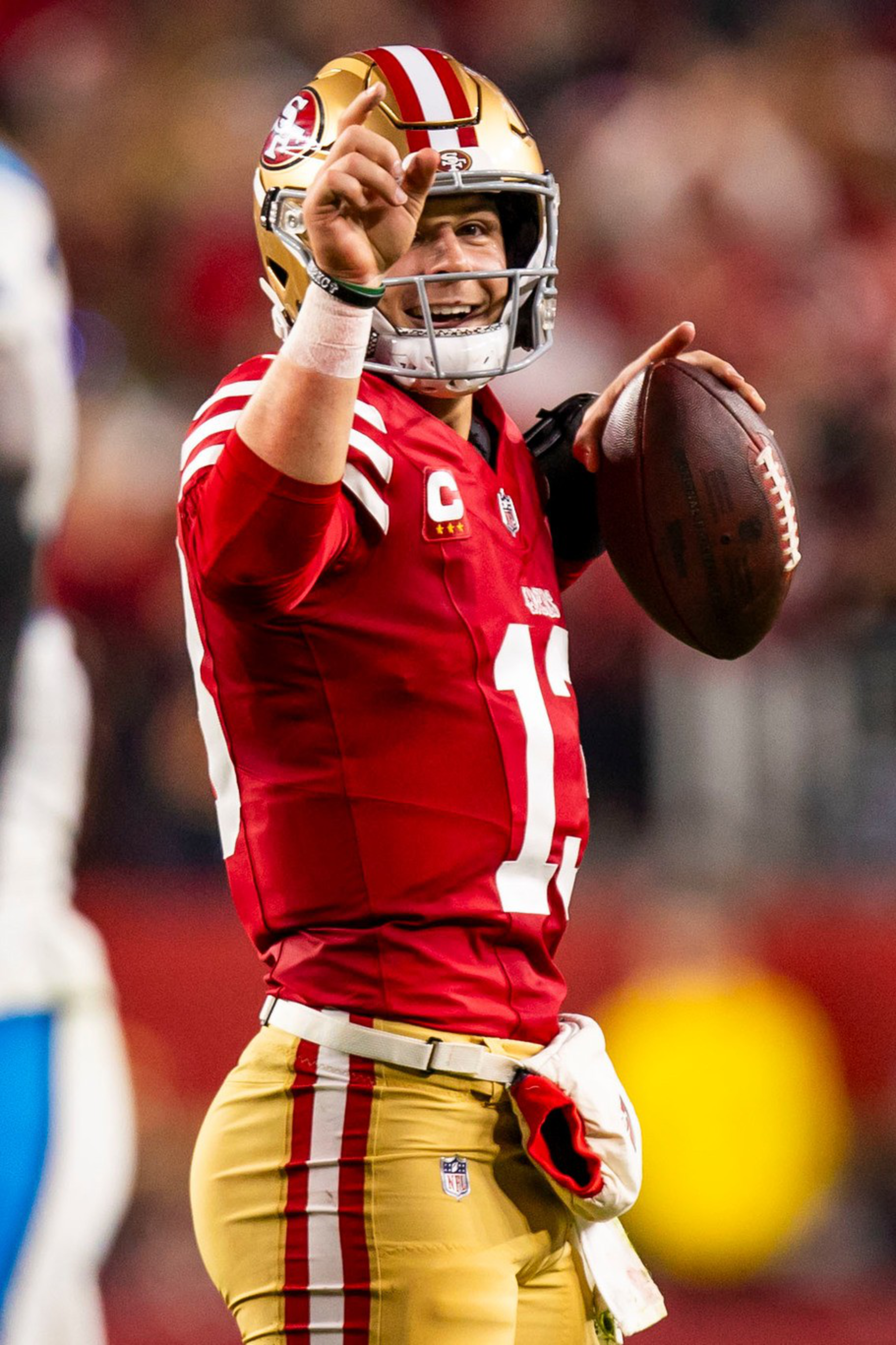 A football player in a red and gold San Francisco 49ers uniform holds a football and points upward with a determined expression during a game.