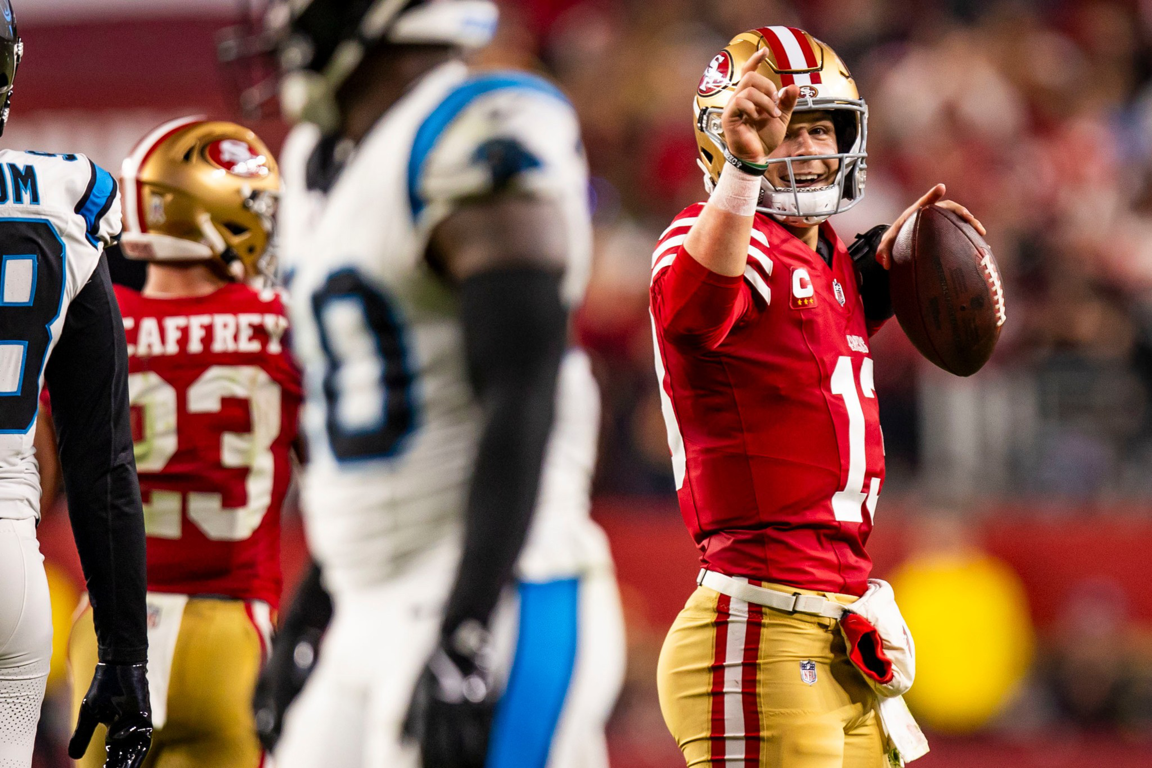 A football player in a red and gold San Francisco 49ers uniform holds a football and points upward with a determined expression during a game.