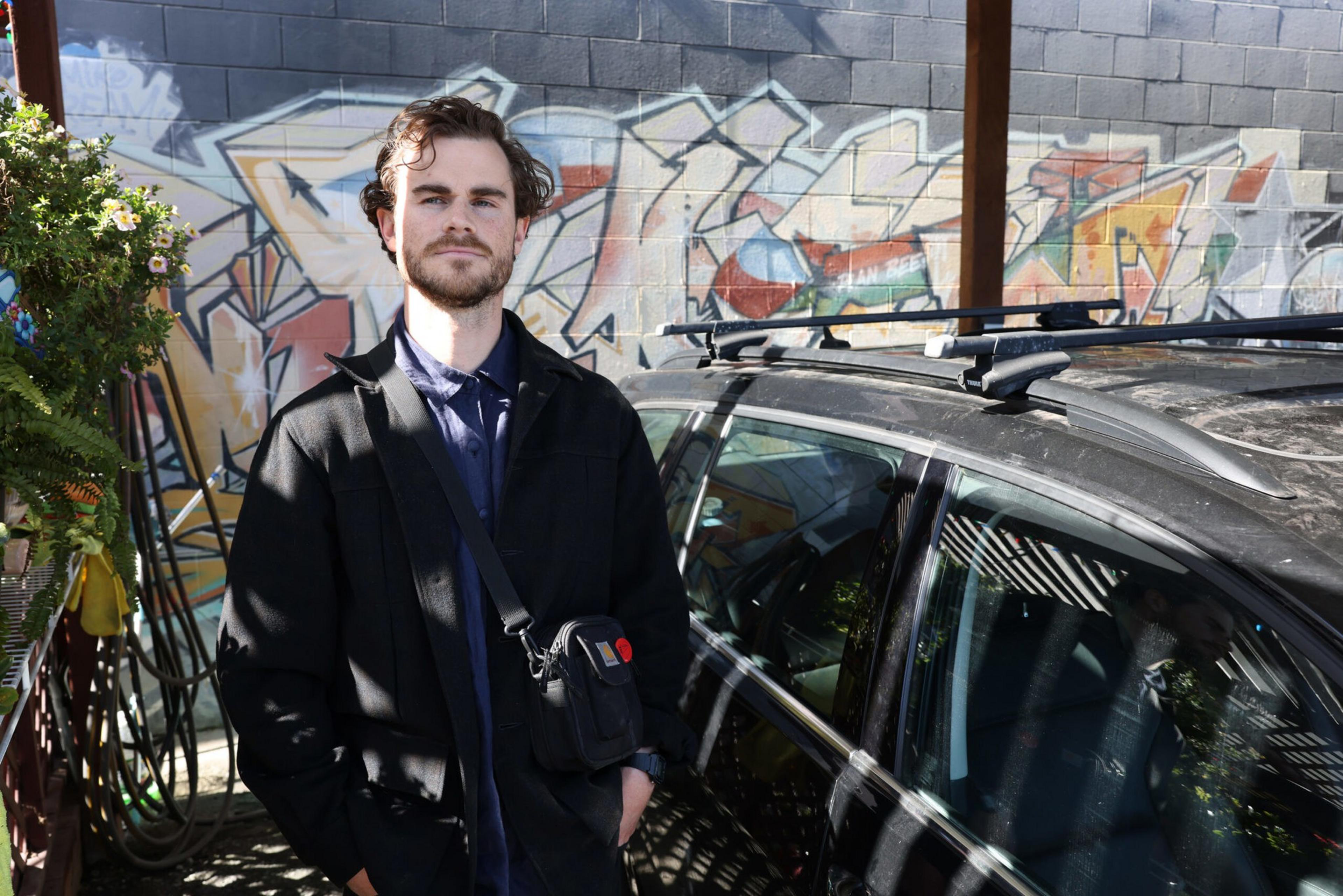 A man with dark hair and beard wearing a black jacket stands next to a black car with a roof rack in front of a graffiti-covered brick wall.