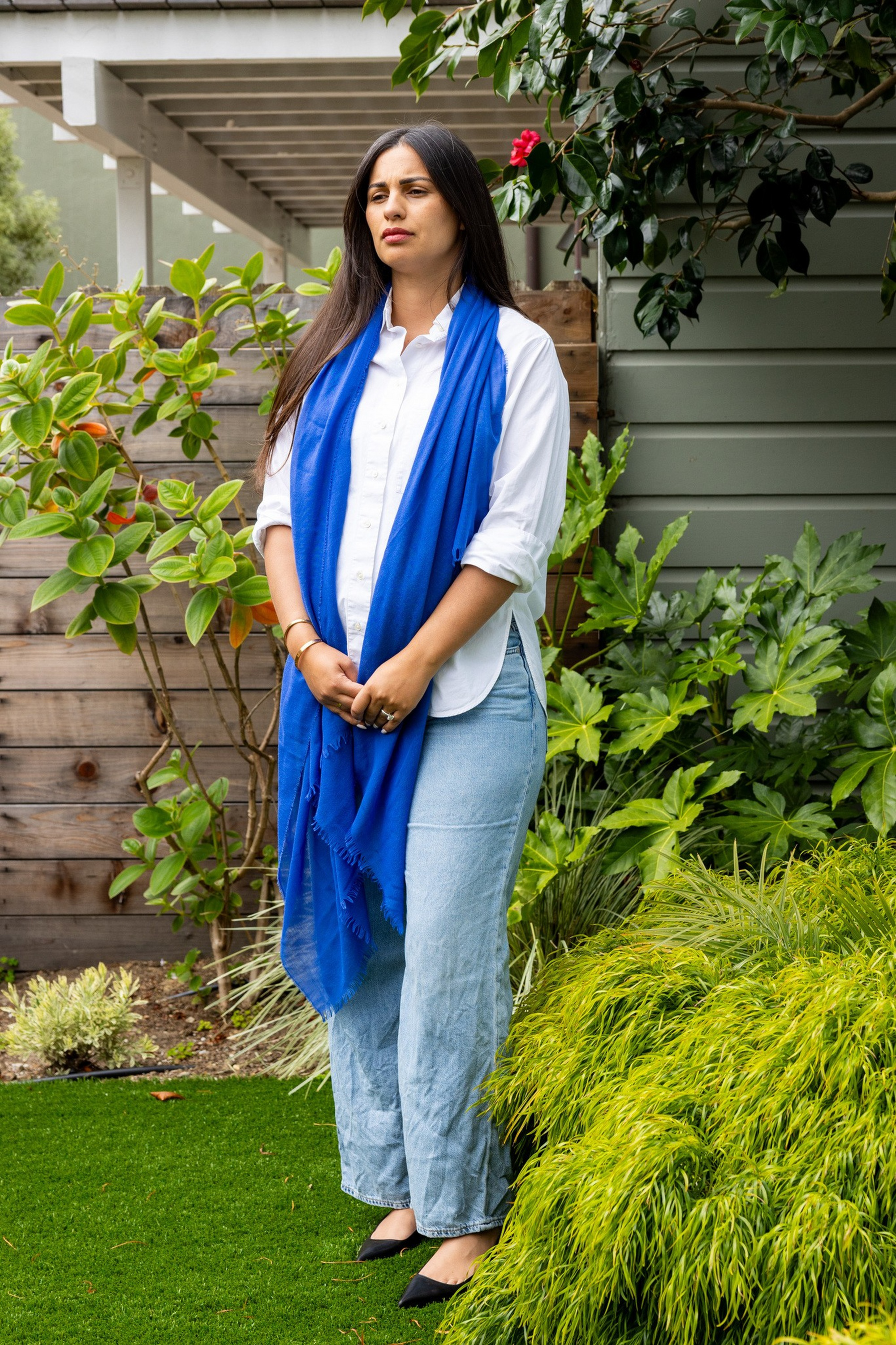 A woman stands in a garden wearing a white shirt, light blue jeans, black shoes, and a bright blue scarf, surrounded by lush green plants.