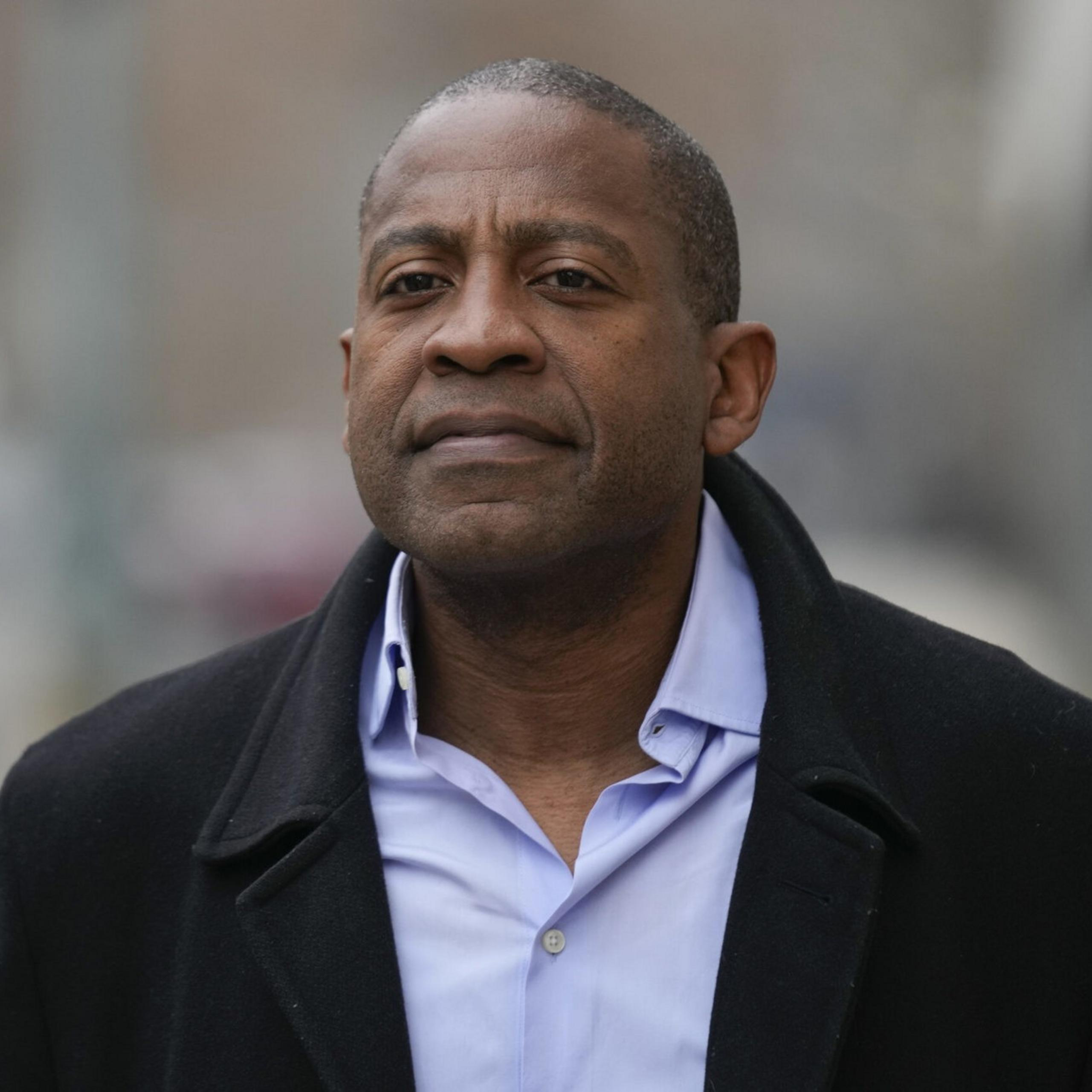 A man is walking outside wearing a dark overcoat and a light blue shirt. The background is blurred, suggesting an urban environment.