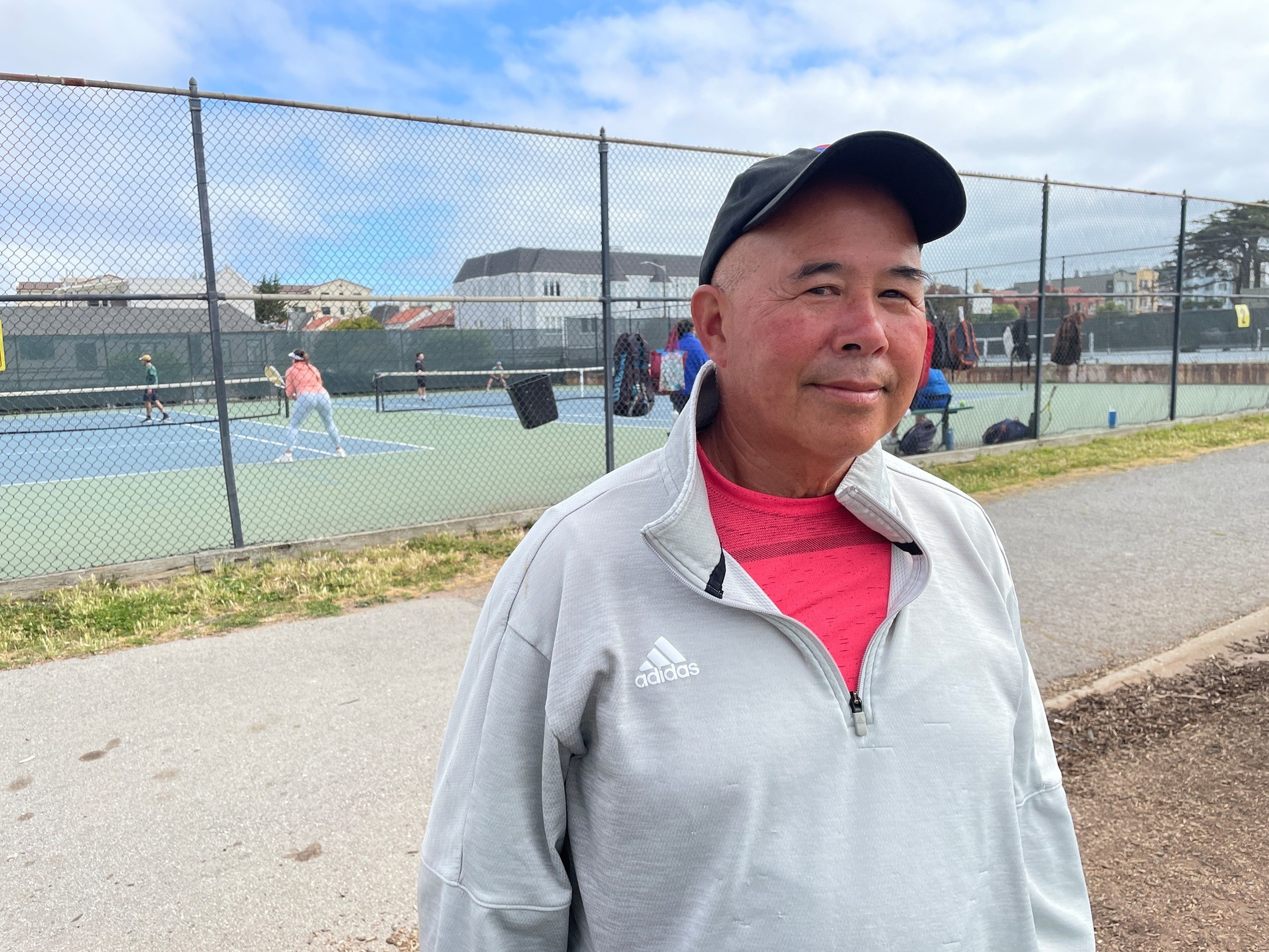 A man wearing a gray Adidas jacket, a red shirt, and a black cap stands near a tennis court with players in action behind a chain-link fence.