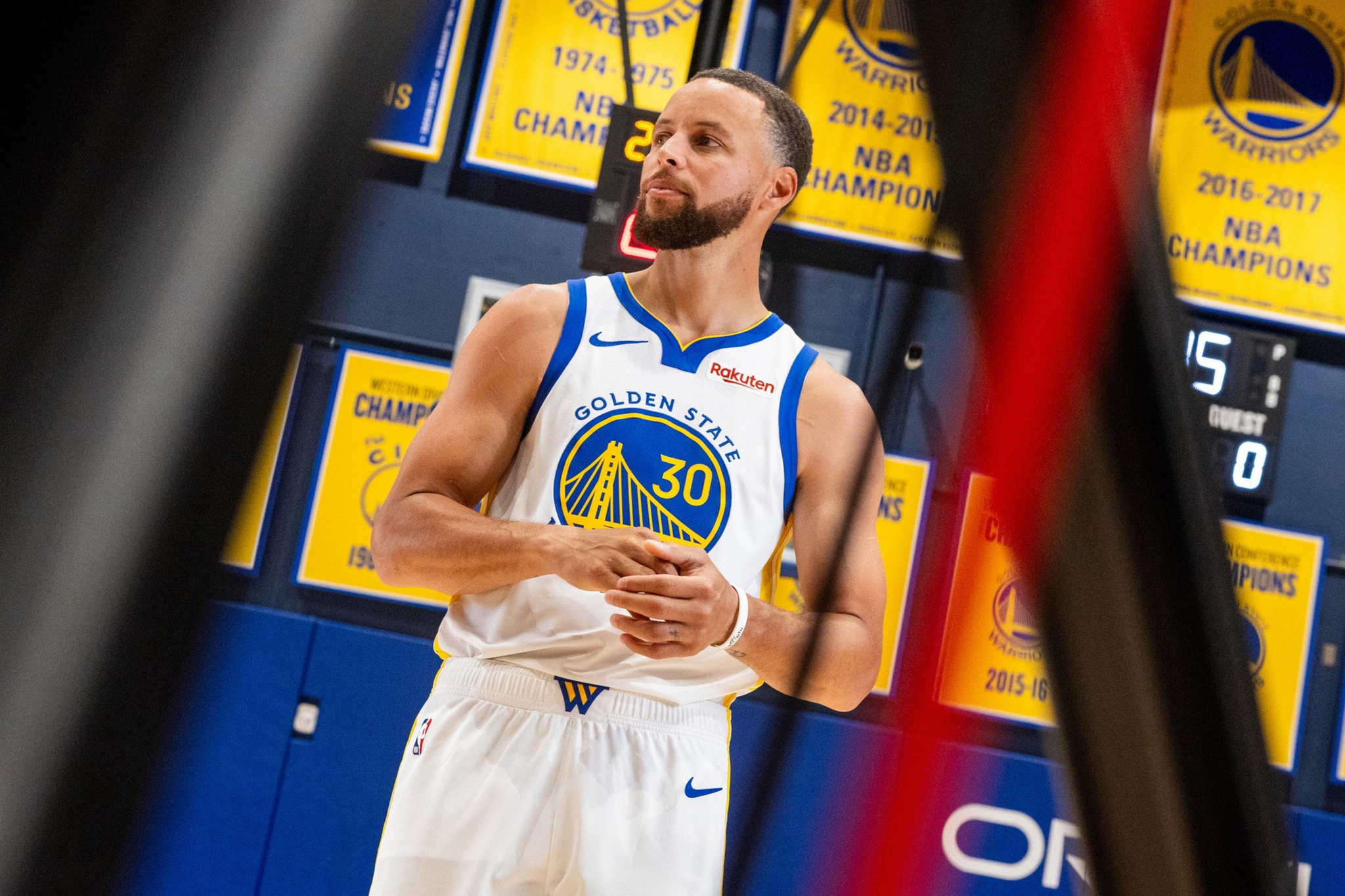 A basketball player in a Golden State Warriors jersey stands in front of championship banners hanging on a gym wall.